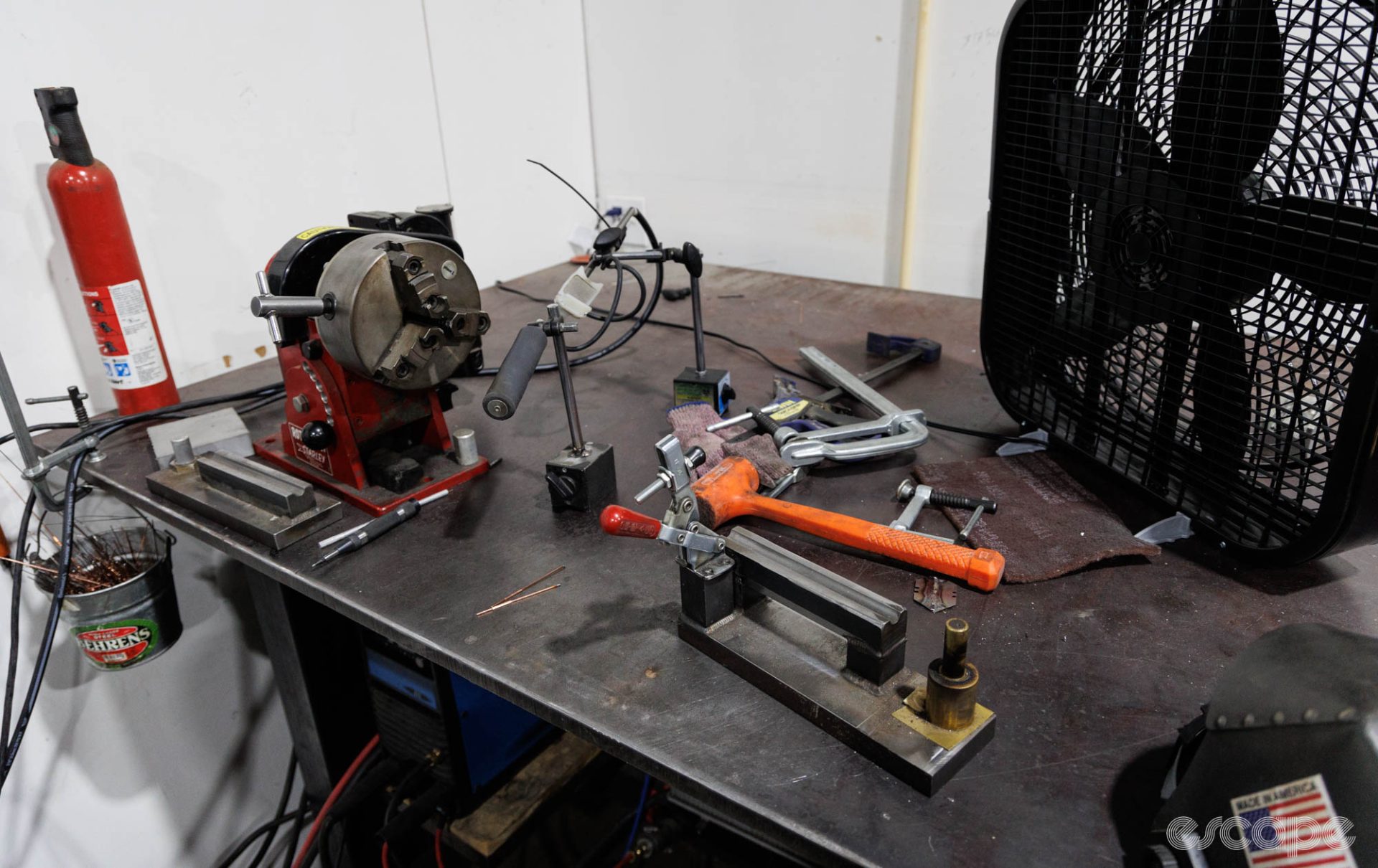 A cluttered bench contains implements for welding tools, with a small bucket of TIG welding rod hanging off one end.