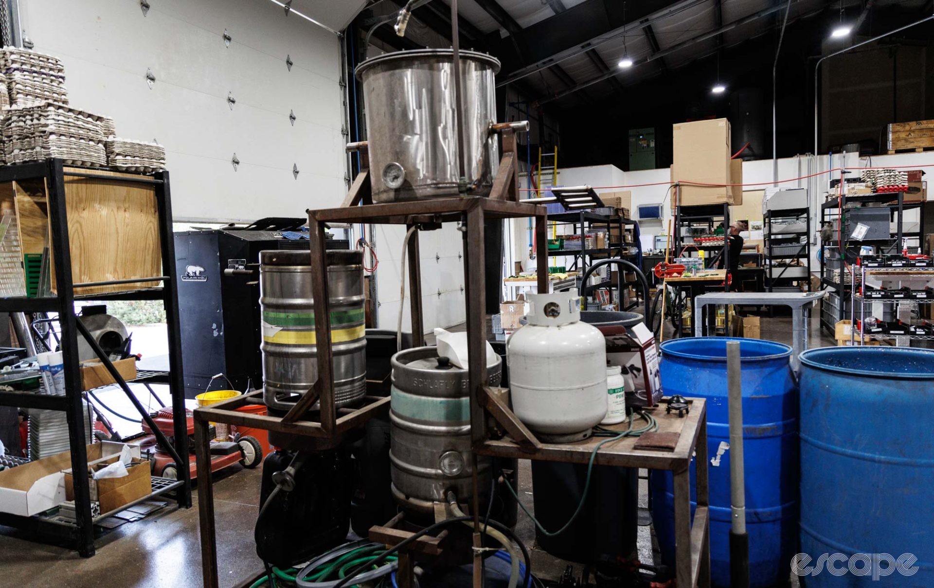 Jason Quade's homebrewing equipment sits on the shop floor. There's a large brewing tun on a high shelf and two kegs below it.