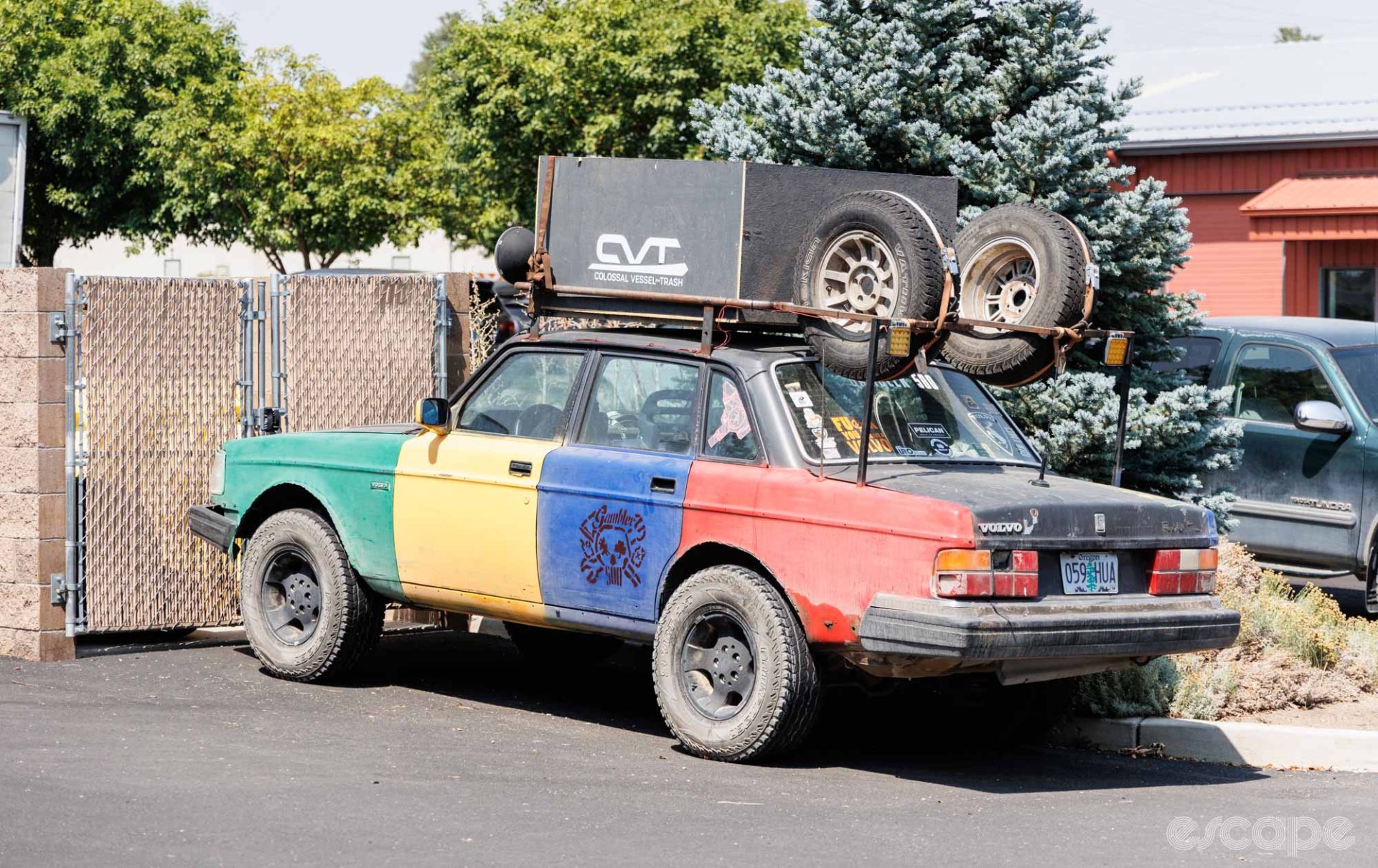 A dilapidated Volvo sedan, with body panels in green, yellow, blue, red, and black. The car is lifted and has oversize rally tires, two spares and a large box on a custom roof rack.
