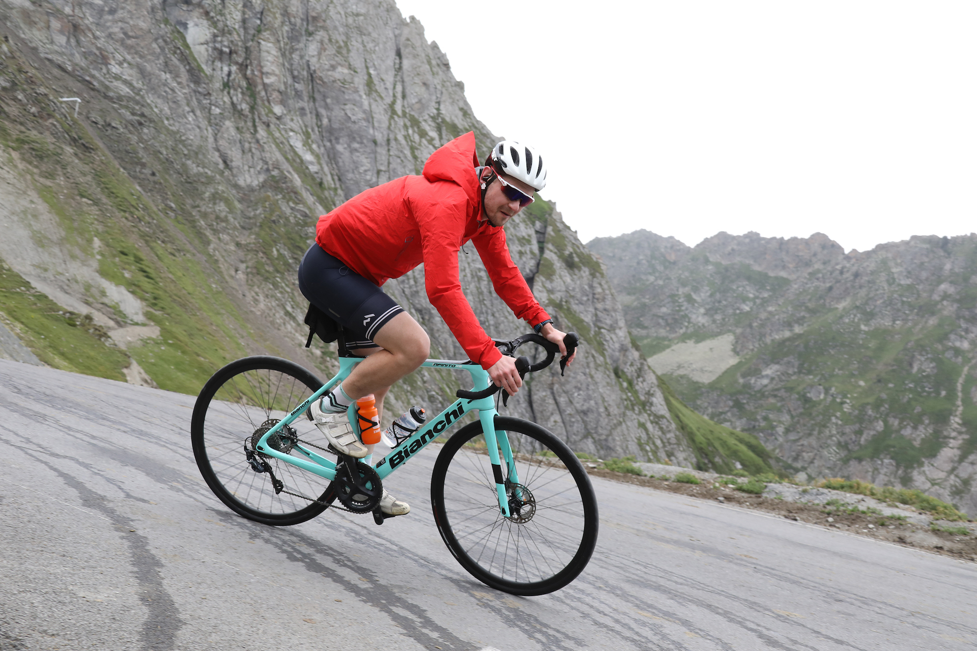 The author on a celeste-green Bianchi Infinito rental bike, wearing black shorts and a red hooded jacket for the chilly descent. He rounds a corner with tall peaks behind him.