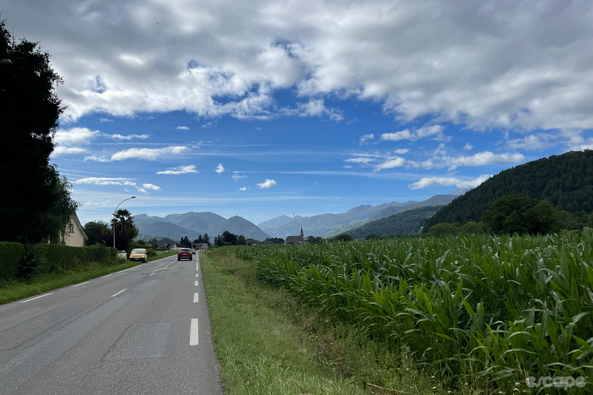 A view of the flat roads leading to the Pyrenees, with fields of corn framed in the distance by tall peaks. A church steeple appears in the middle distance.