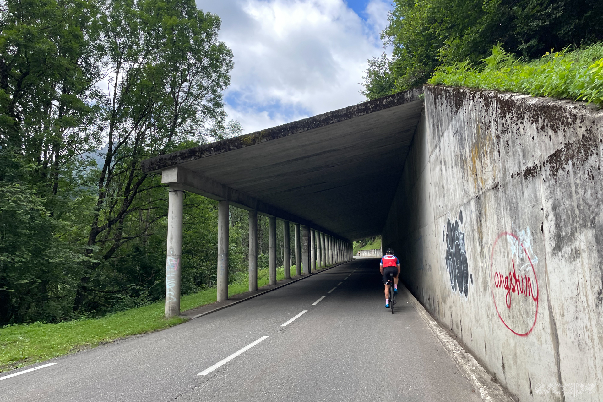 A cyclist enters a tunnel built for avalanche control on the climb up the Tourmalet. There is a tall concrete wall to the uphill side supporting a roof, and the downhill side is open, with only support columns.