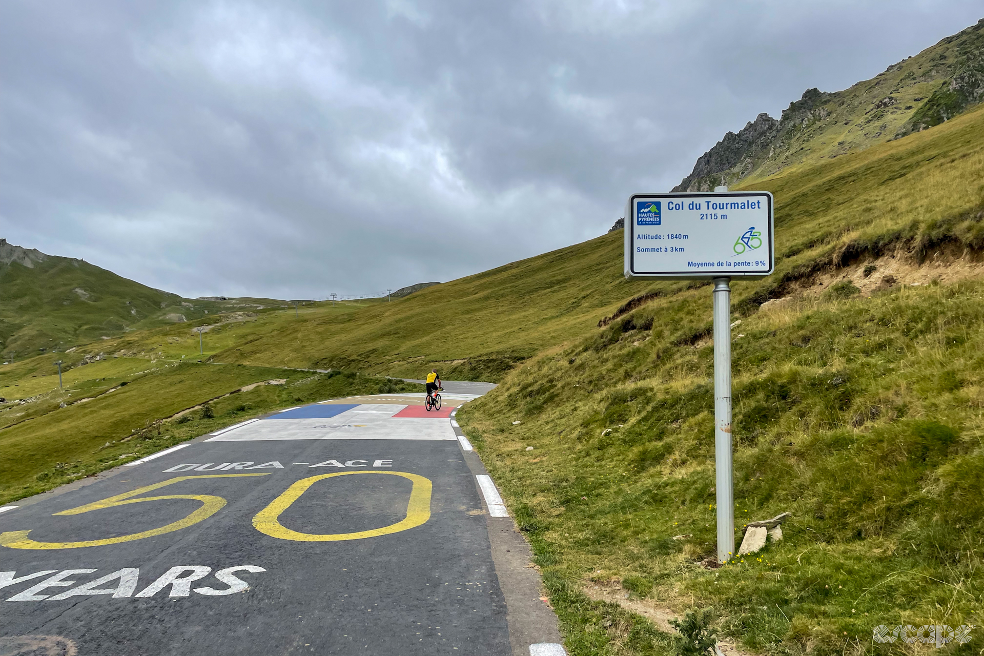 High on the Col du Tourmalet, there's a sign announcing the peak and summit (2,115 meters) and current altitude (1,849 meters) with 3 km to go to the summit, at a 9 percent gradient. The road is painted with the colors of the French flag and a corporate marketing message for the 50th anniversary of Shimano's Dura-Ace components.