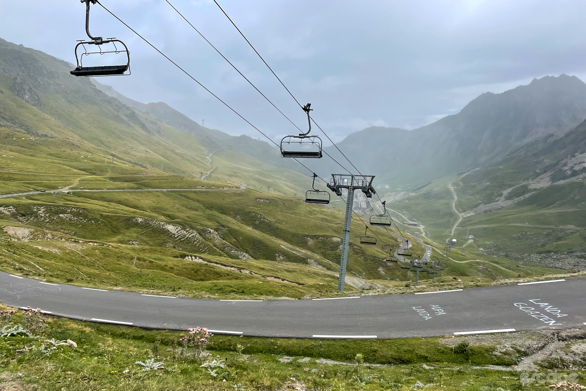 A dormant ski lift crosses the road up the Tourmalet, with empty chairs against a misty, green landscape.