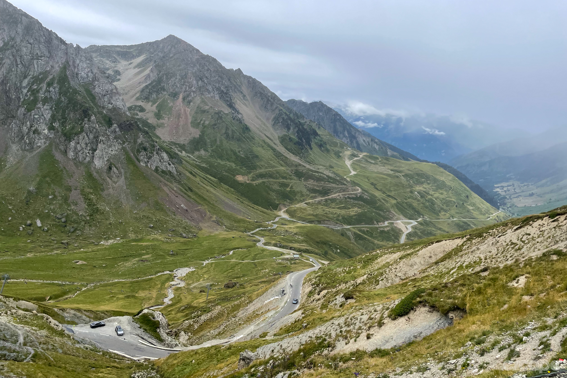 The view down valley from the summit of the Col du Tourmalet: a ribbon of pavement winds down the hillside, with spurs branching off at odd intervals. On either side large peaks rise up, and the summits are covered in clouds.