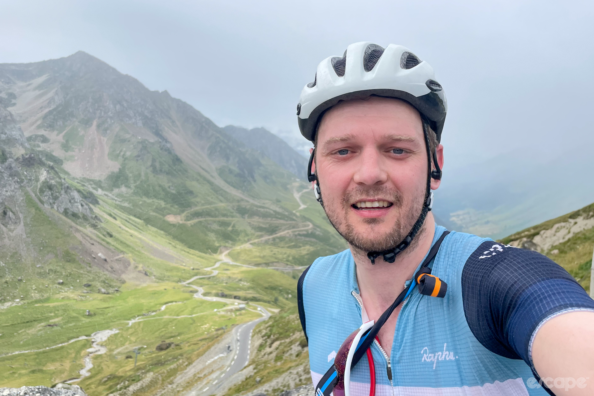 The author on the summit of the Tourmalet, wearing a jersey with light and dark blue blocks and a white helmet. Behind him is a road off the Tourmalet, heading down valley.