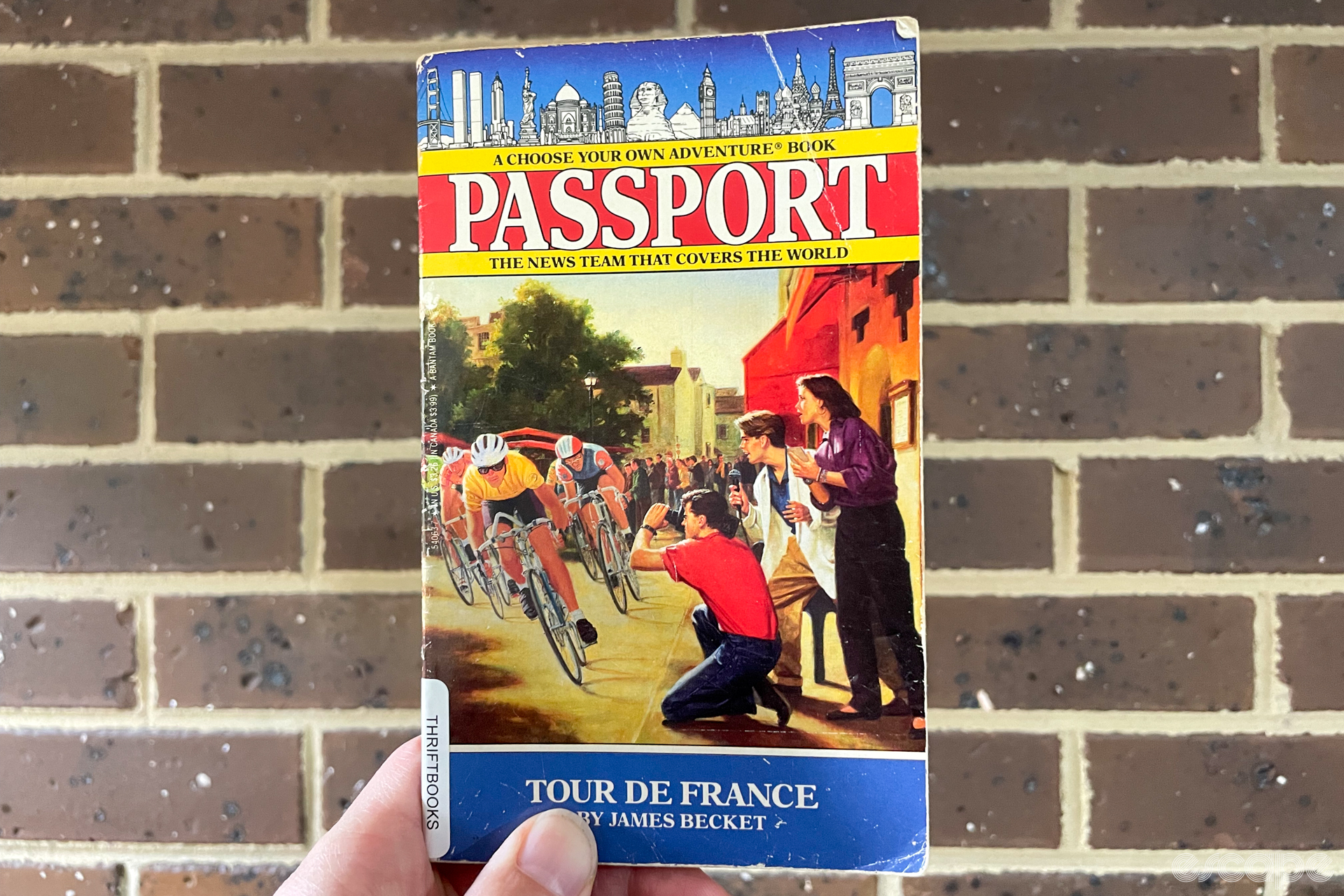 A photo of the cover of a Tour de France-themed Choose Your Own Adventure book in the 'Passport' series, written by James Becket. The book is being held by someone's left hand, with several fingers visible, and with a brick wall in the background.