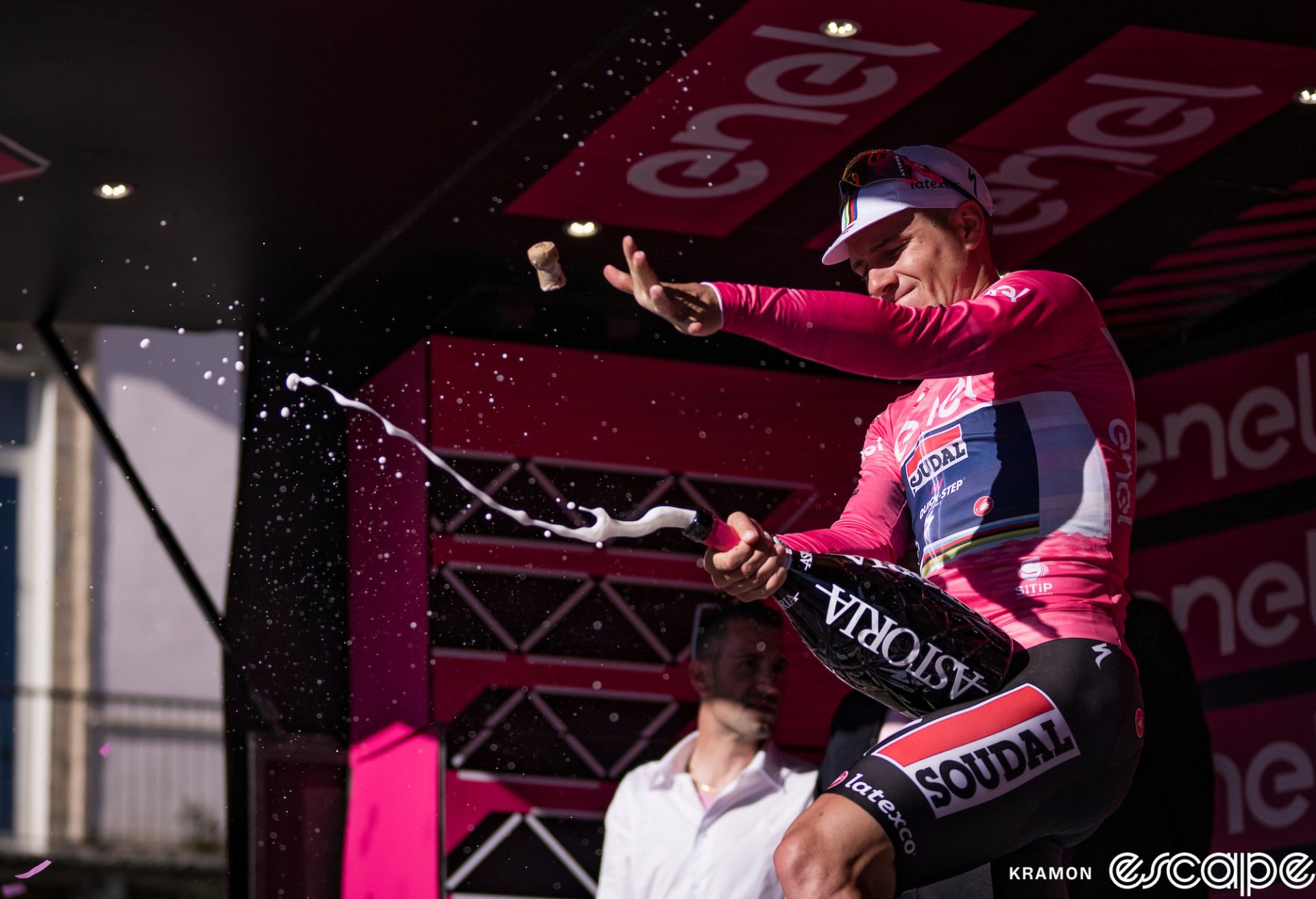 Remco Evenepoel cracks a giant bottle of champagne on the podium of stage 1 of the 2023 Giro d'Italia. He's in the pink jersey of race leader, and the cork is flying through the air in front of his hand as champagne starts to bubble and flow out of the bottle.