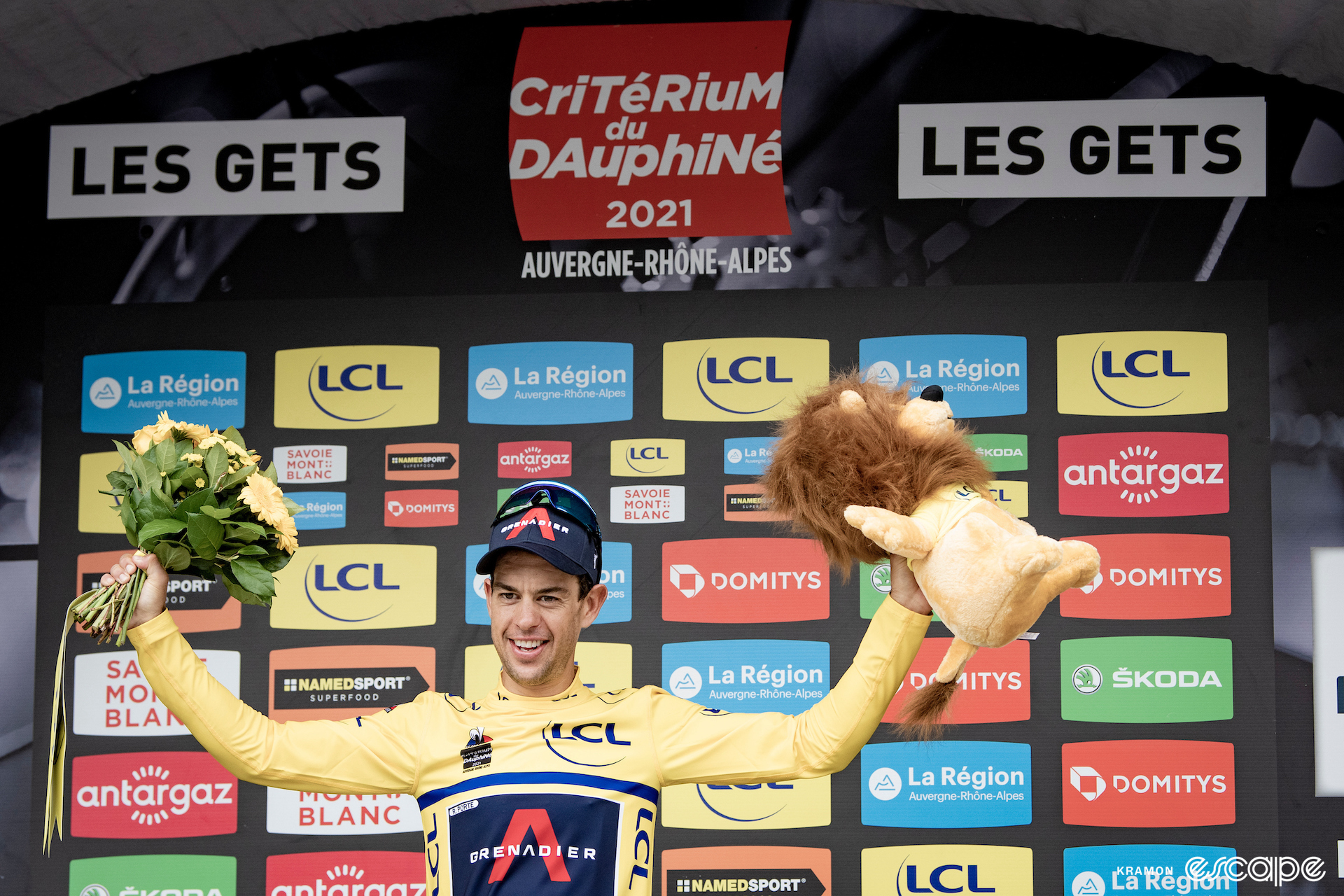 Professional cyclist Richie Porte stands on the podium at the 2021 Critérium du Dauphiné, wearing the yellow leader's jersey, holding a yellow lion in his left hand and a bunch of flowers in his right hand.
