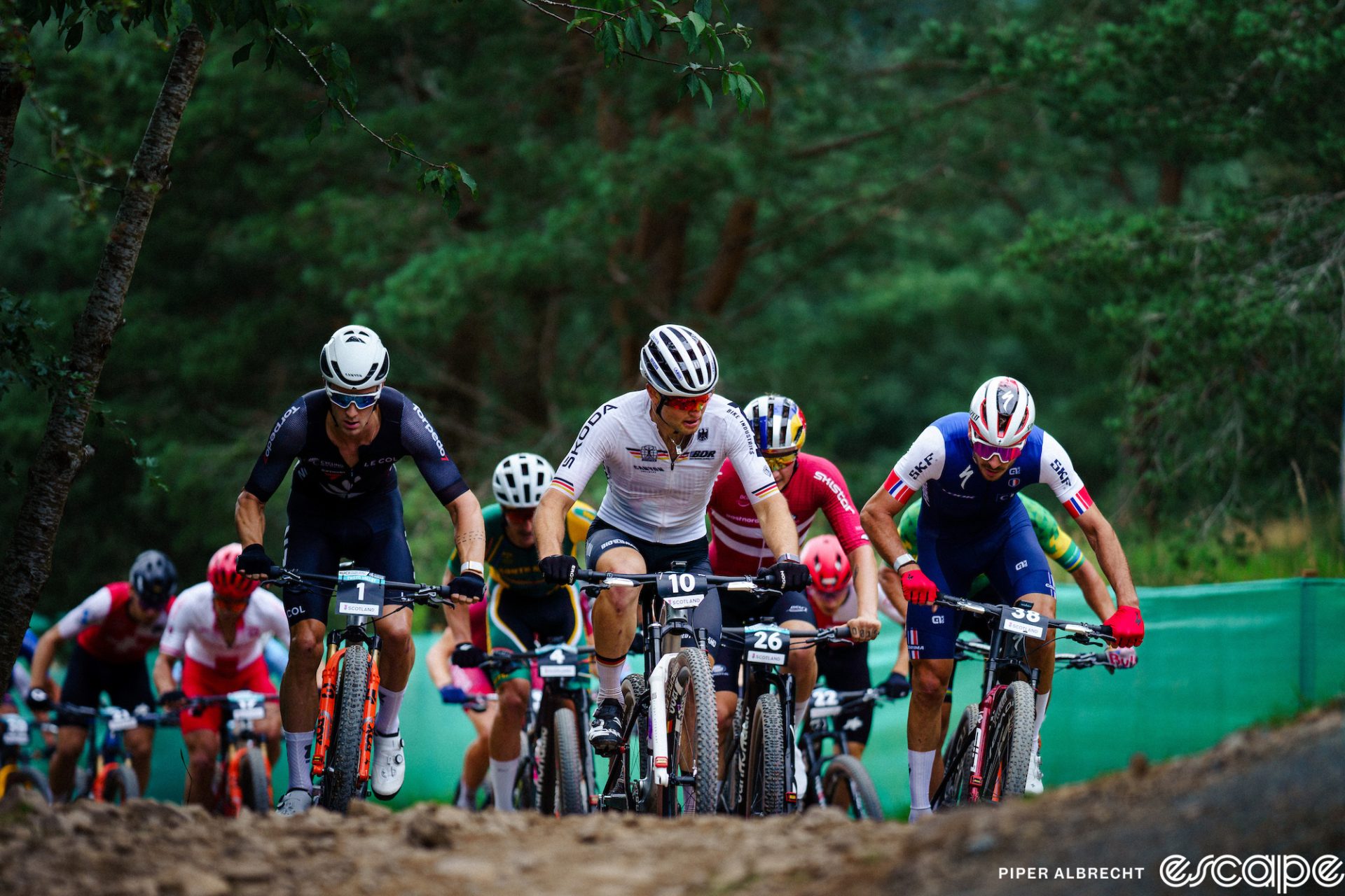 From left to right, Sam Gaze, Luca Schwarzbauer, and Victor Koretzky lead the men's field in the short track at the 2023 World MTB Championships. Koretzky is out of the saddle and Schwarzbauer is looking to his left as if to mark Koretzky's move. Gaze is looking straight ahead as the pack rides up a short climb.
