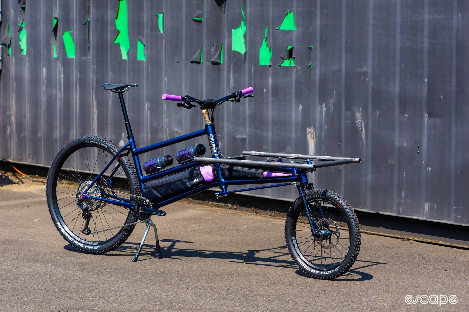 Photo showing two-wheel cargo bike from Schön Studio, with platform mounted at the front. It is a glossy metallic blue with pink highlights. 