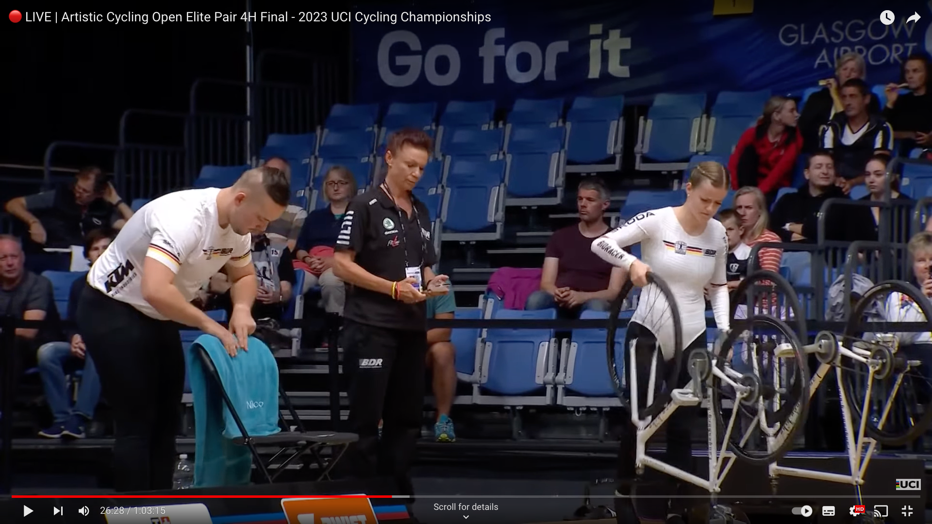 Nico Rödiger and Leah Styber prepare to begin their routine in the elite pairs final. Rödiger is placing a towel with a 'Nico' monogram on his chair on the courtside, carefully smoothing out wrinkles. 