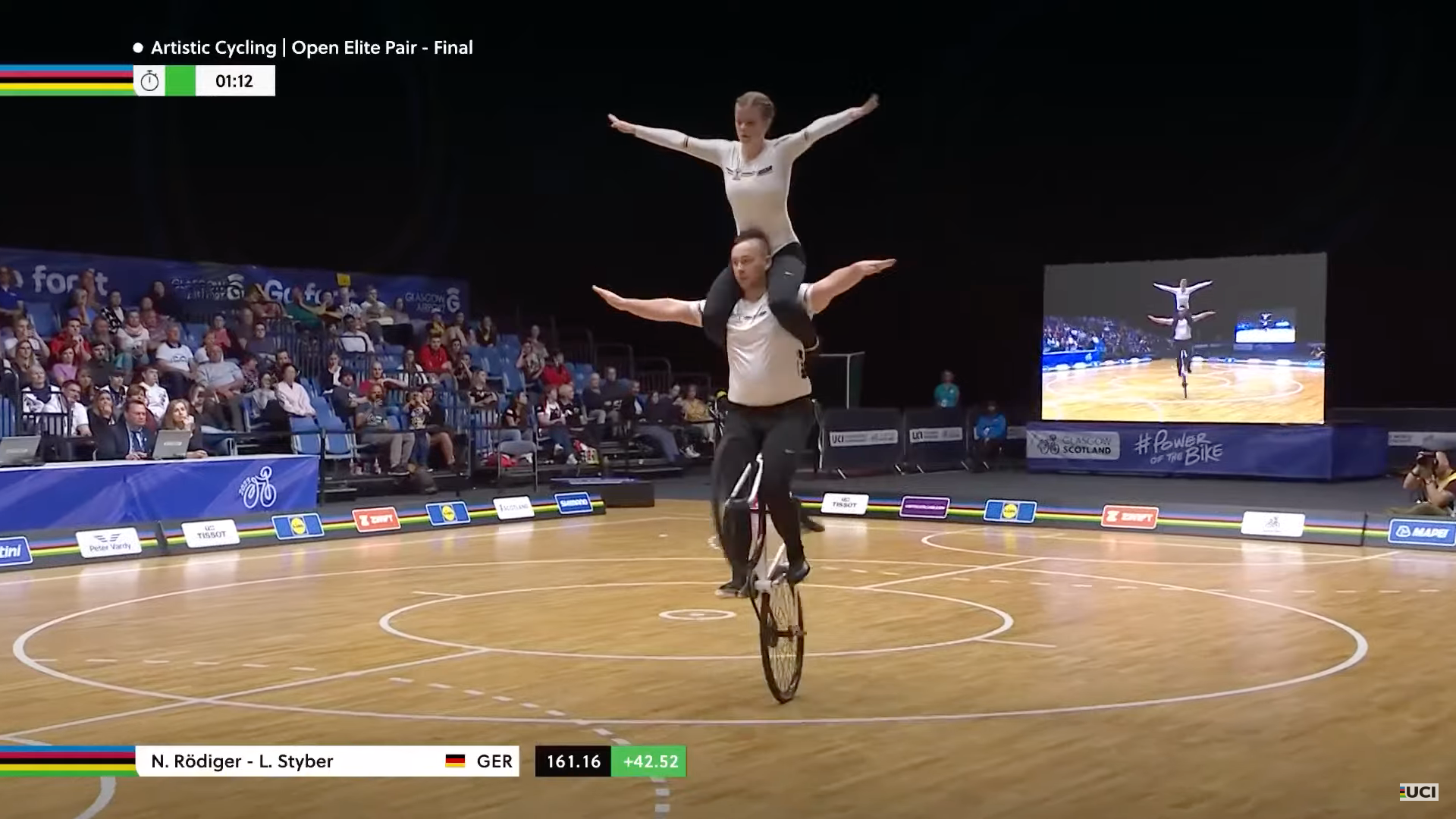 Youtube screenshot of a German artistic cycling pair. They are both on a single bike with the  female rider sitting on the male rider's shoulders while the bike is doing a wheelie.