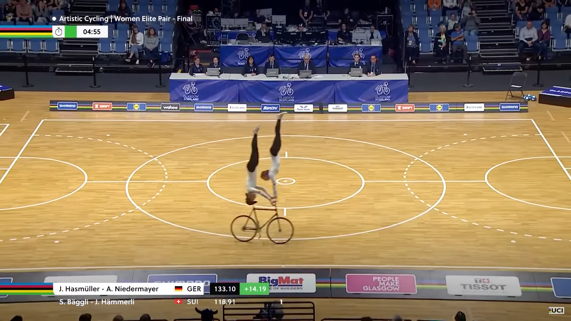 Two cyclists in the women's elite pairs final, both of them on the same bike. One is doing a handstand on the handlebars, the other is doing a headstand on the saddle.