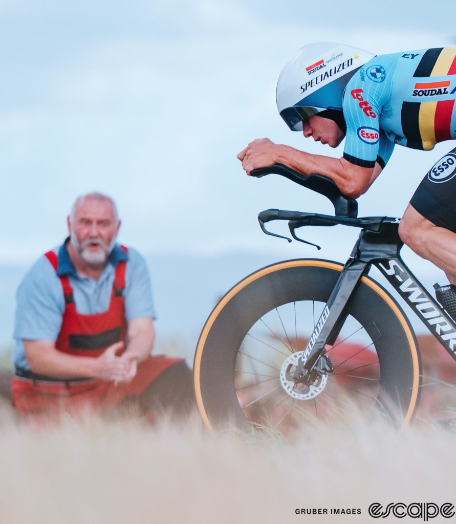 Evenepoel at TT Worlds. He's shown in profile, but only his front half, in an aero tuck in full concentration as he rides by a slightly out-of-focus bearded Scottish man dressed in red overalls and a blue shirt.