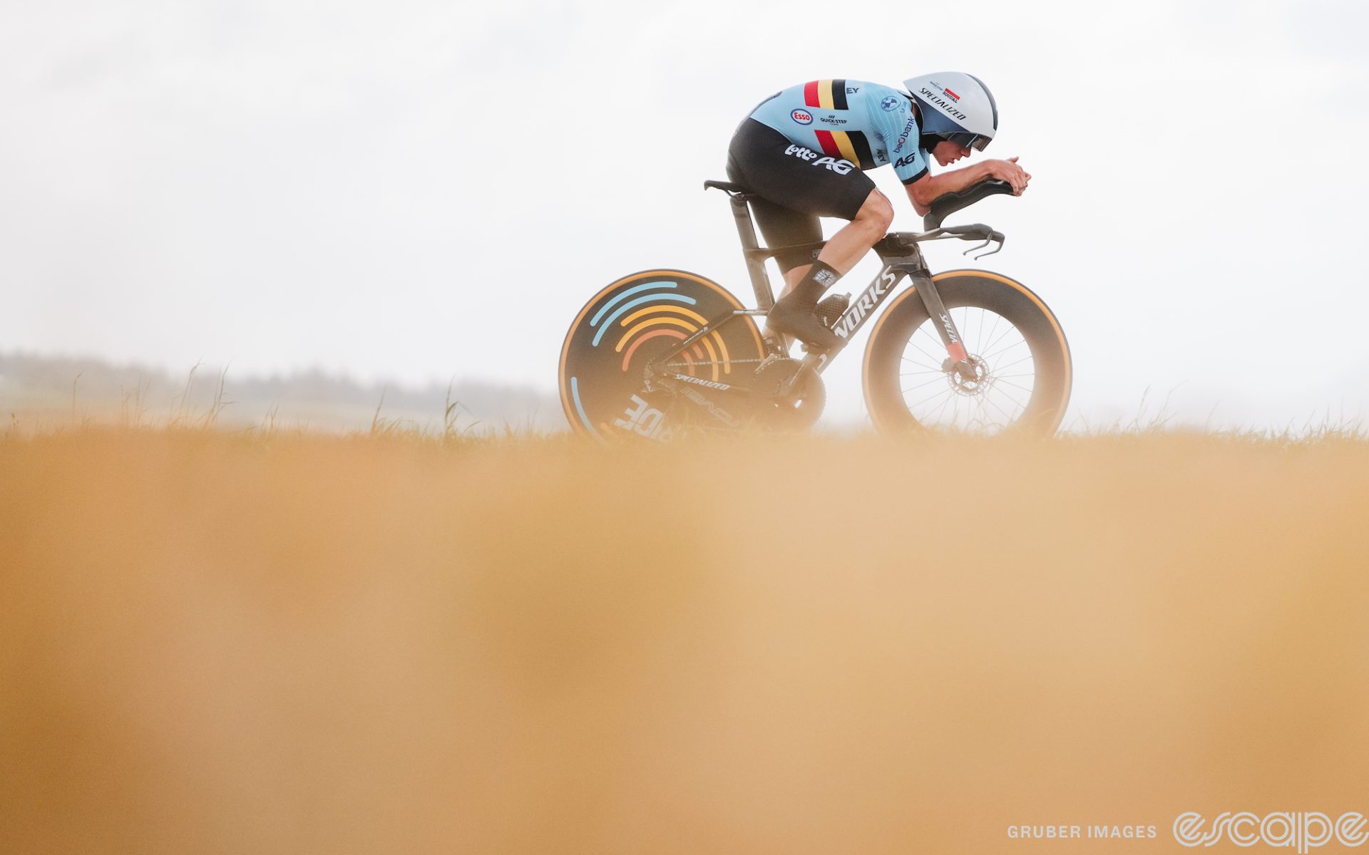 Remco Evenepoel races in the World Time Trial Championship in Glasgow, Scotland. The picture is divided in roughly two equal halves. The top is Evenepoel, in Belgian national team kit, in an efficient aero tuck on his time trial bike with a grey sky and low hill in the background. The lower half is the blurred-out beige of a field, giving a dreamy kind of feel to the image.