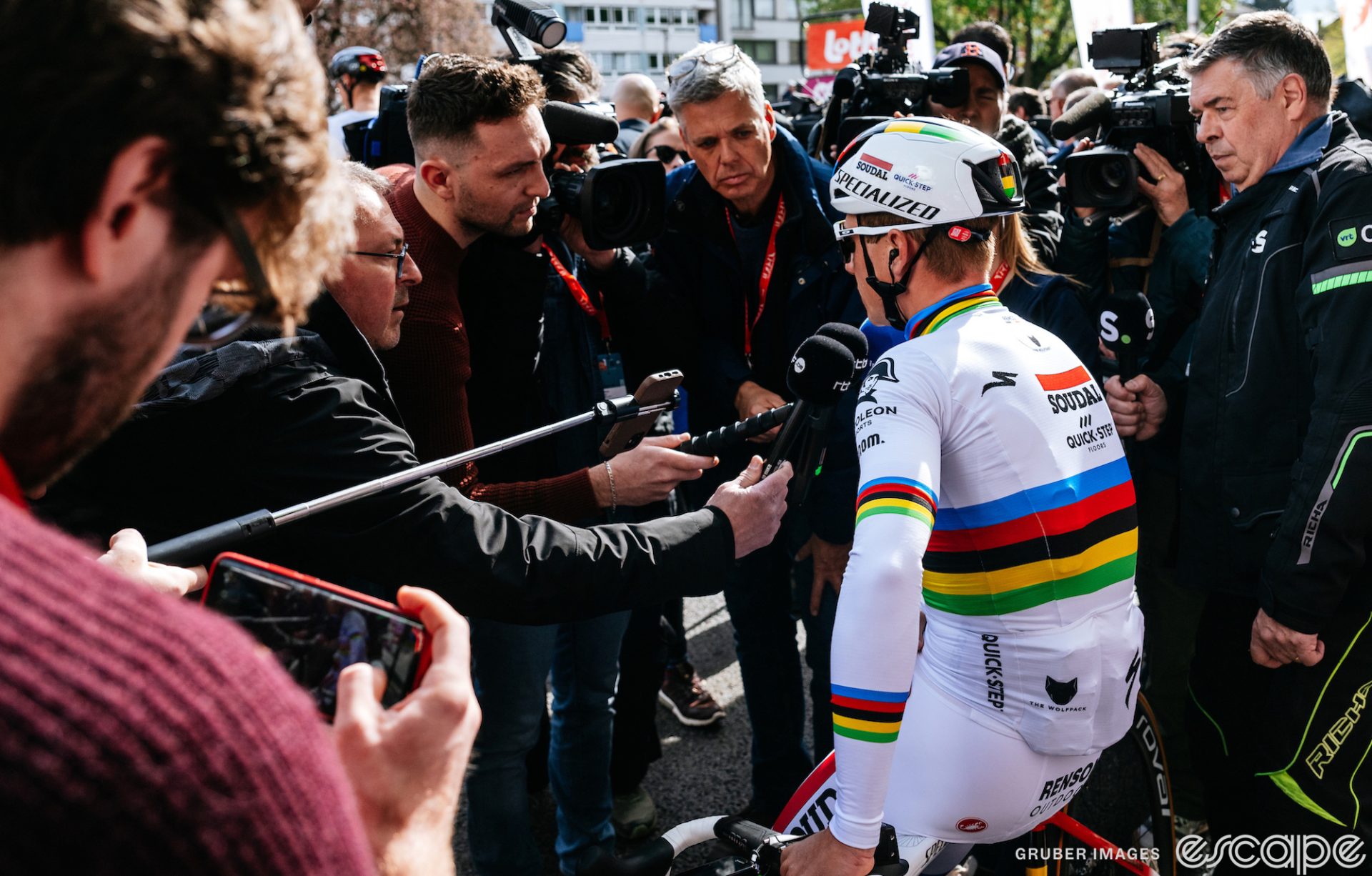 World Champion Remco Evenepoel faces a group of reporters at the 2023 Liege-Bastogne-Liege. Evenepoel is sitting on his bicycle's top tube, facing slightly away from the camera, with a bunch of microphones in his face.