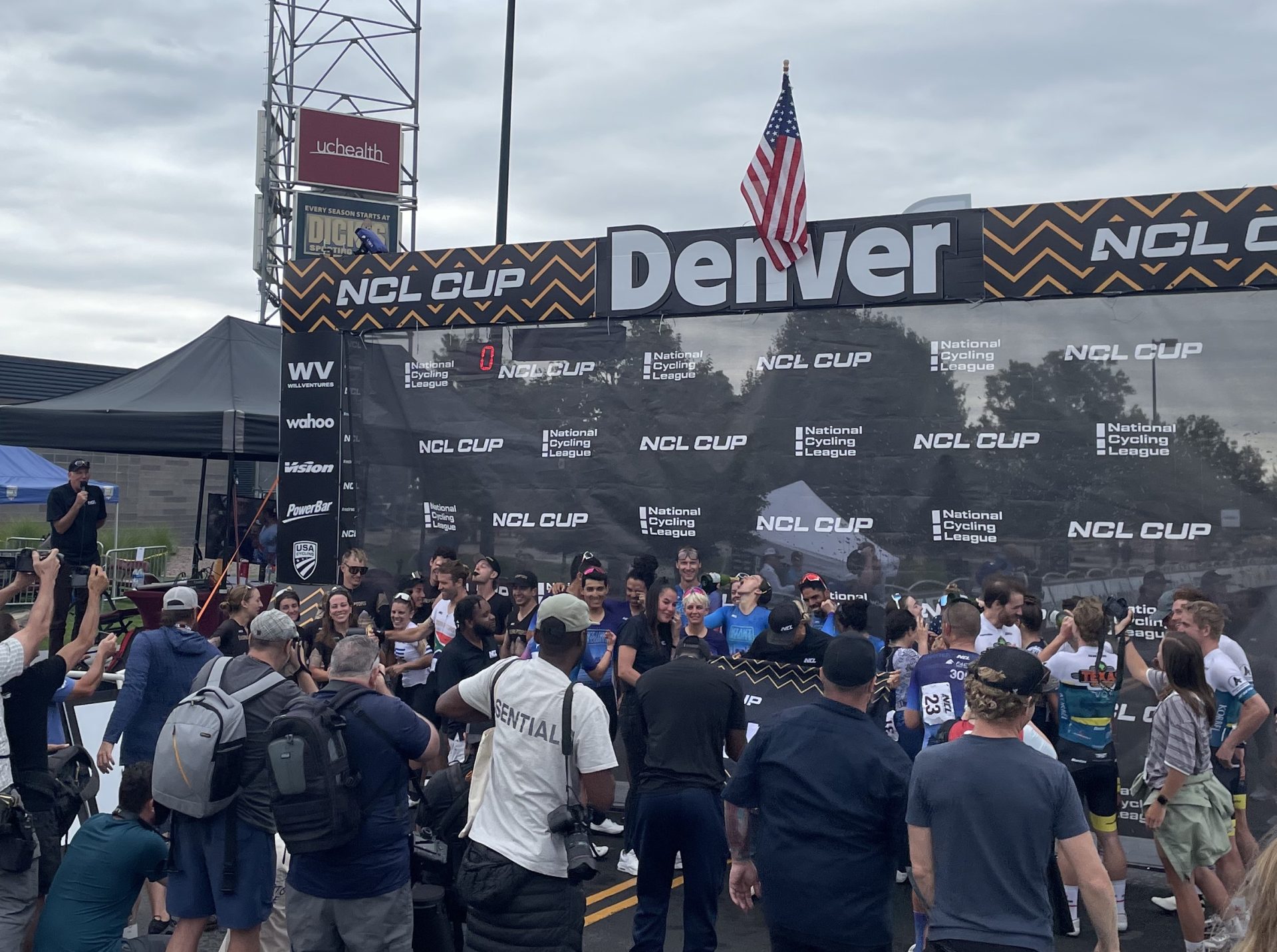 The podium area at the Denver round of the NCL Cup features a line of racers from the Denver Disruptors, Texas Roadhouse-Goldman Sachs, and winning Miami Nights teams. They're surrounded by a few dozen spectators. In the background, race announcer Dave Towle emcees.