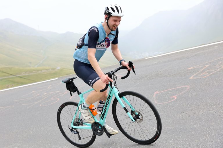 A cyclist climbing the Col du Tourmalet on a celeste-green Bianchi Infinito bike, dressed in black shorts and a light blue CyclingTips jersey with dark blue shoulders and a white helmet. The road tilts precipitously, and the peaks behind him are slightly obscured by mist.