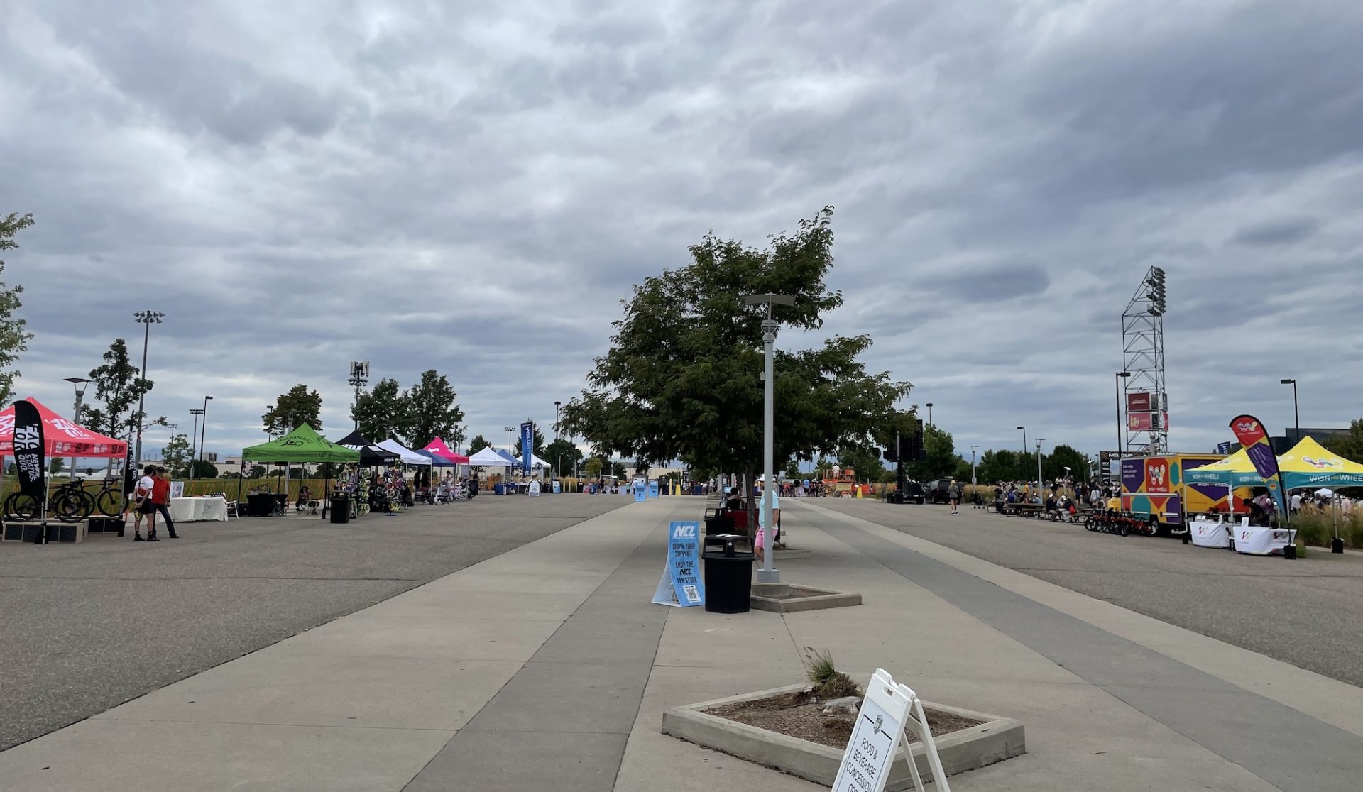 The fan zone expo at the Denver NCL Cup featured two rows of pop-up sponsor tents along a paved area with a central landscape section of trees. There are very few fans present, and the expo area is almost entirely empty of people.