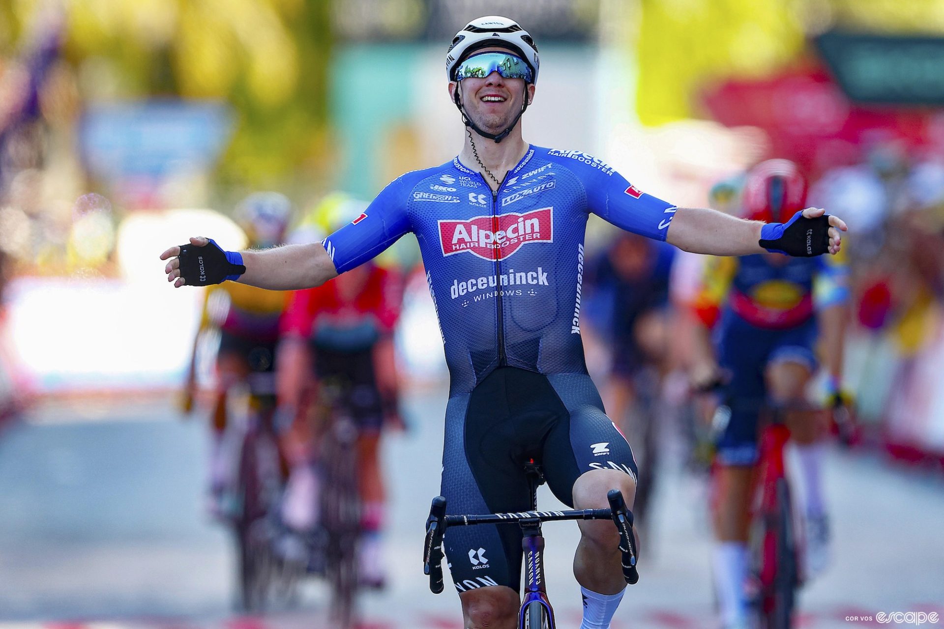 Professional cyclist Kaden Groves, dressed in the primarily dark blue kit of Alpecin-Deceuninck, celebrates a stage victory by extending his arms horizontally.