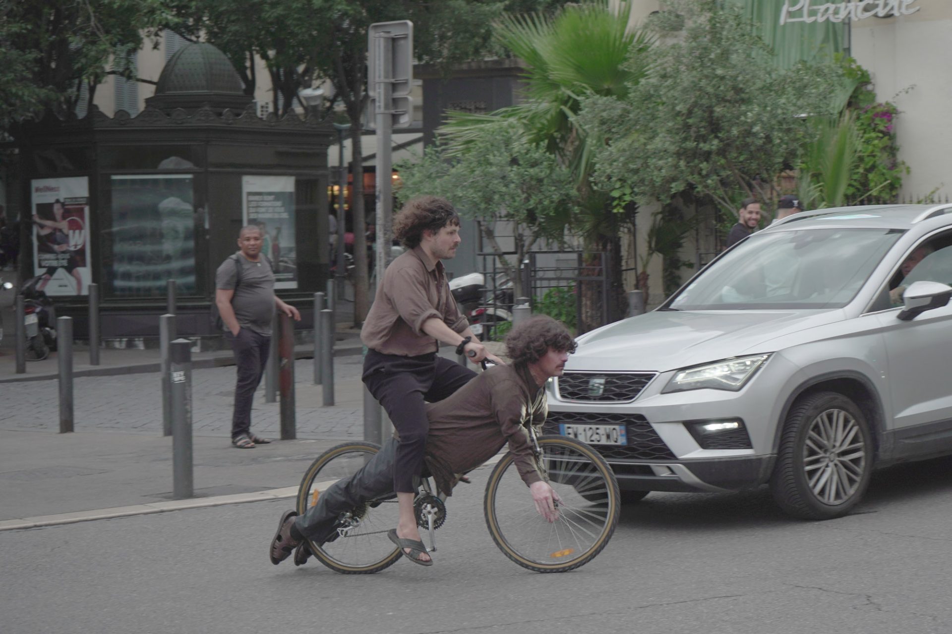 Barzic rides his bicycle through traffic. Two onlookers in the background watch with visible amusement. 