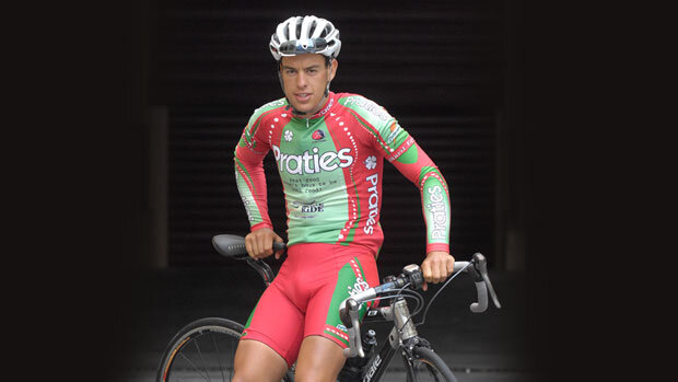 Professional cyclist Richie Porte leans on the top tube of a Cannondale road bike in 2008. He's dressed in the red and green kit of the Praties cycling team, he has a white helmet on with sunglasses stuck through the vents.