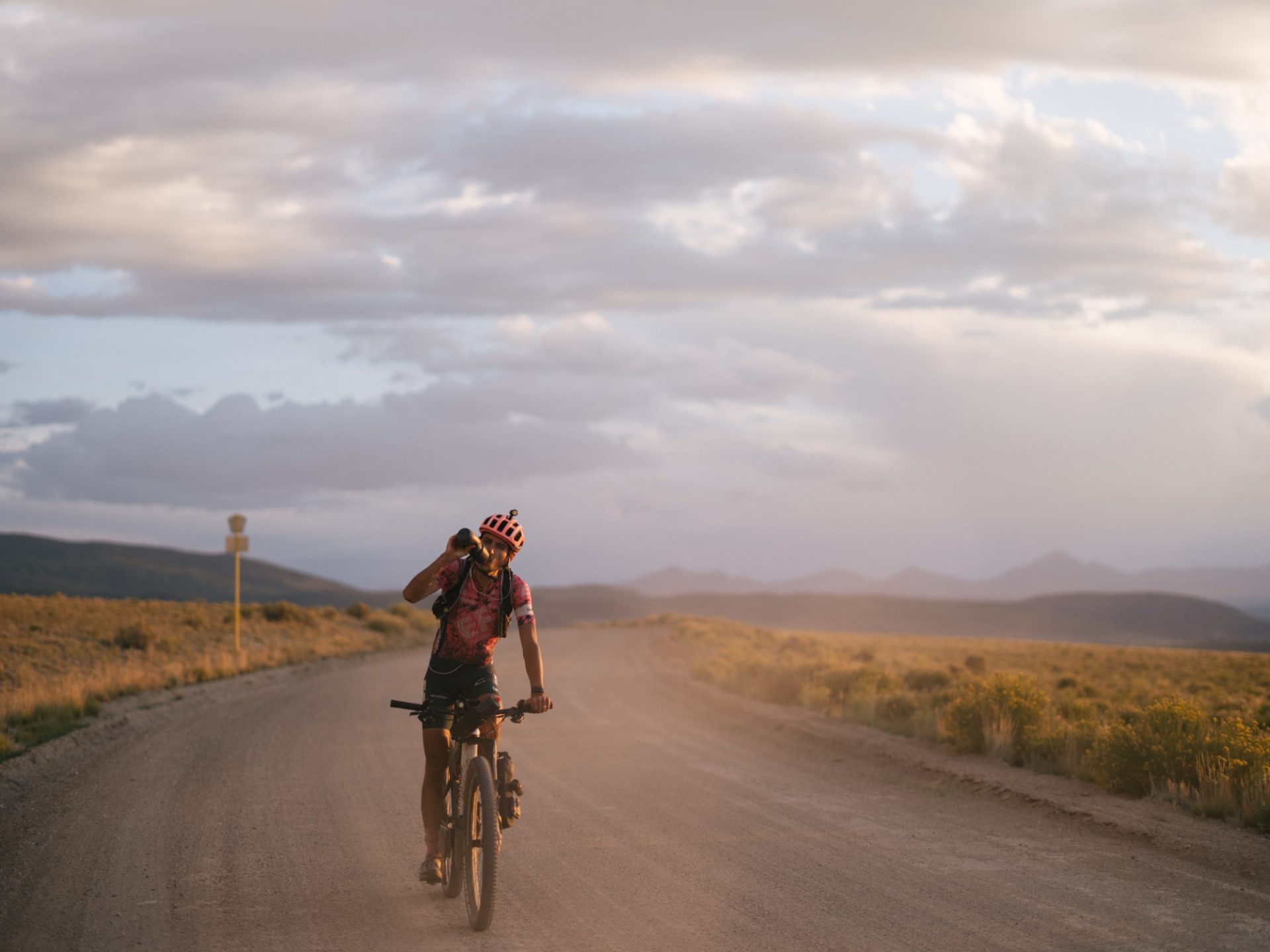 Lachlan Morton drinks from a water bottle while riding his mountain bike on a dirt road at dusk.