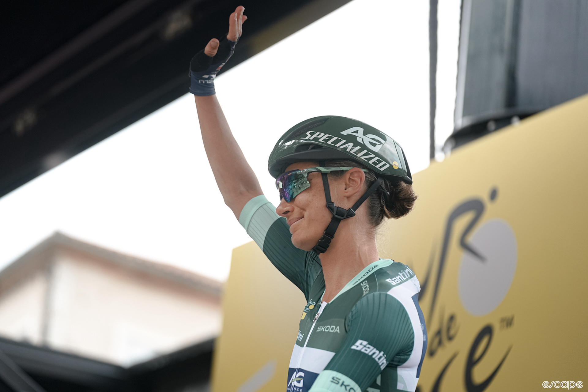Ashleigh Moolman Pasio waves to the crowd at the start of stage 4 of the Tour de France Femmes. She's wearing the green jersey of the leader of the points competition, with a matching green helmet.