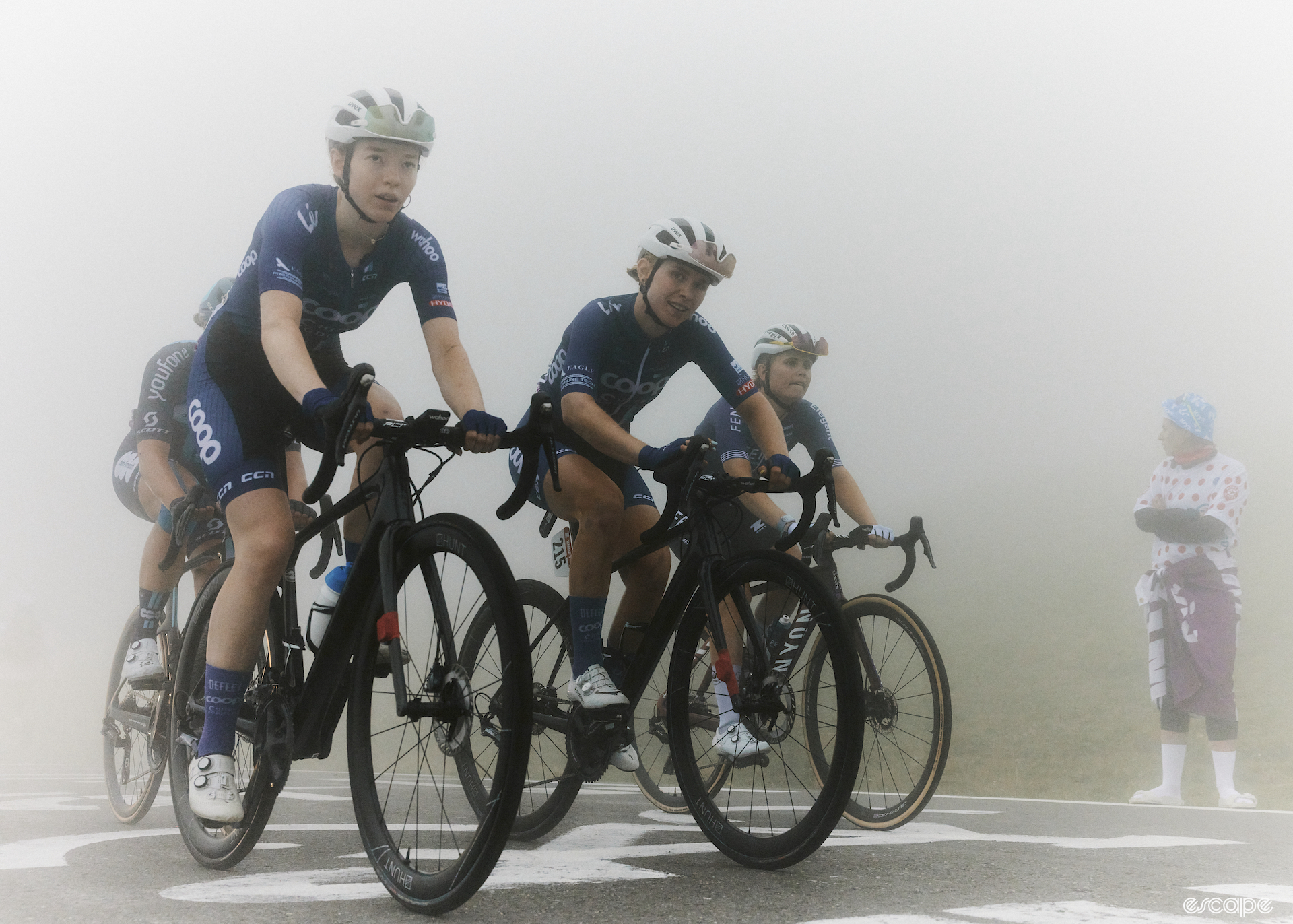 Three riders of the same team ride side by side up a mountain, surrounded by mist. 