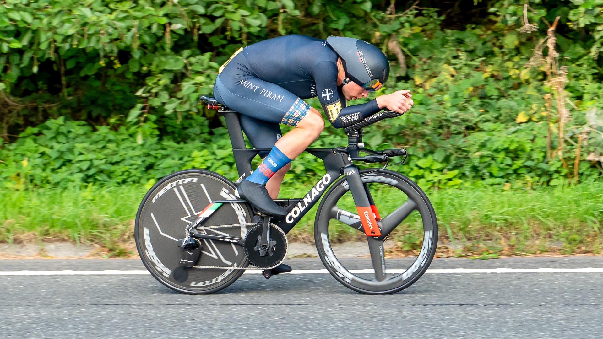 The image shows Josh Charlton of St Piran riding a Colnago time trial bike with an Endura TT helmet. Charlton is riding in the time trial position and is clearly moving at speed. The background is overgrown hedge or ditch reaching right up to the roads edge.