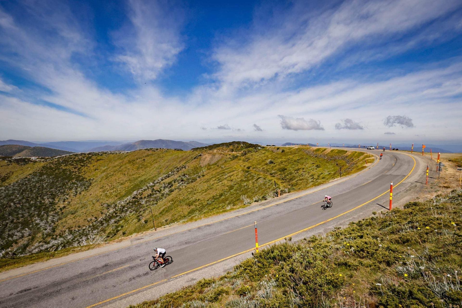 A pair of cyclists, separated by around 30 metres, descend off the summit of Mt. Hotham in the Victorian Alps under blue, cloudy skies.