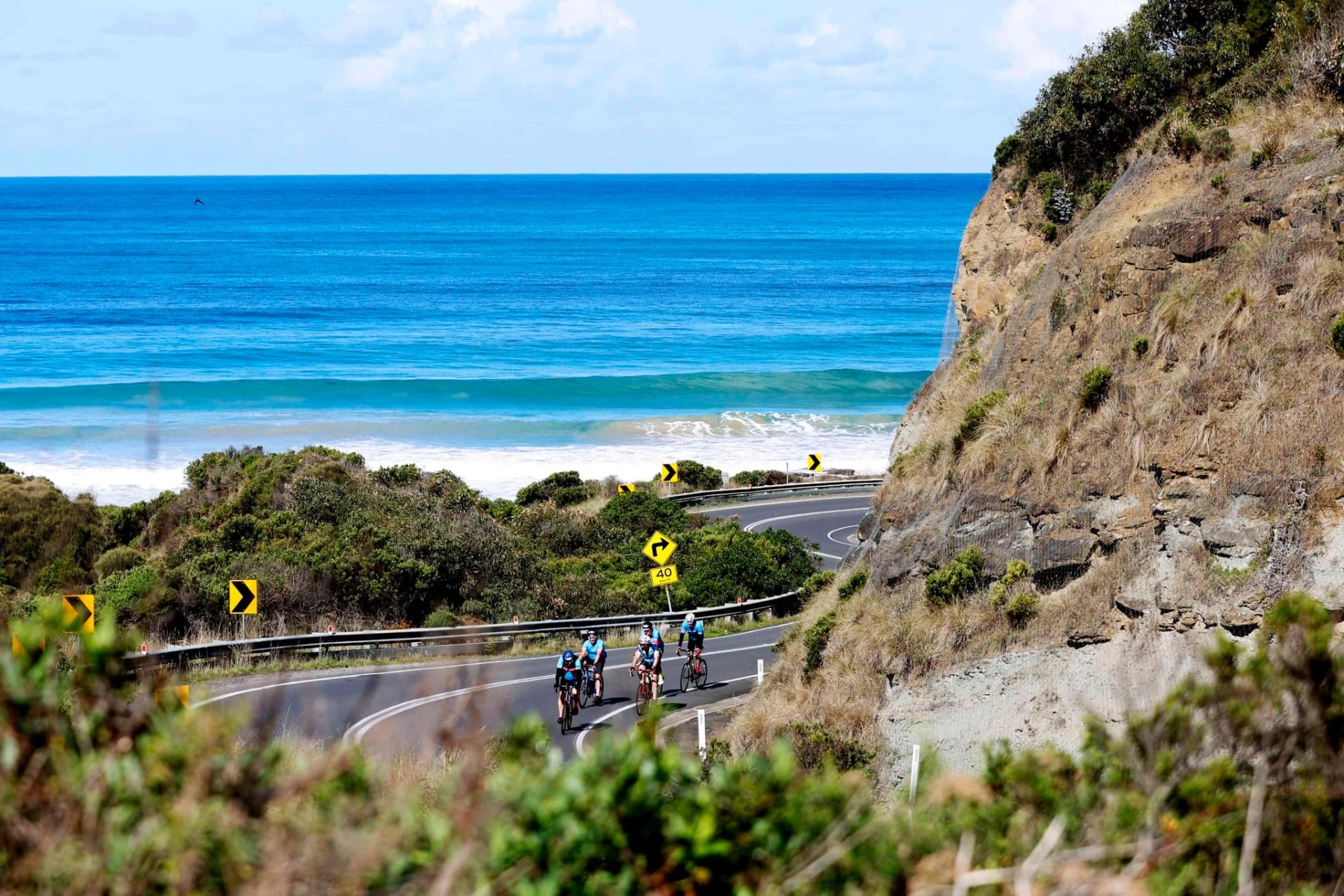 A small group of cyclist rides along the Great Ocean Road, a coastal route in Victoria, Australia. The road hugs the cliffs with the ocean in the background.