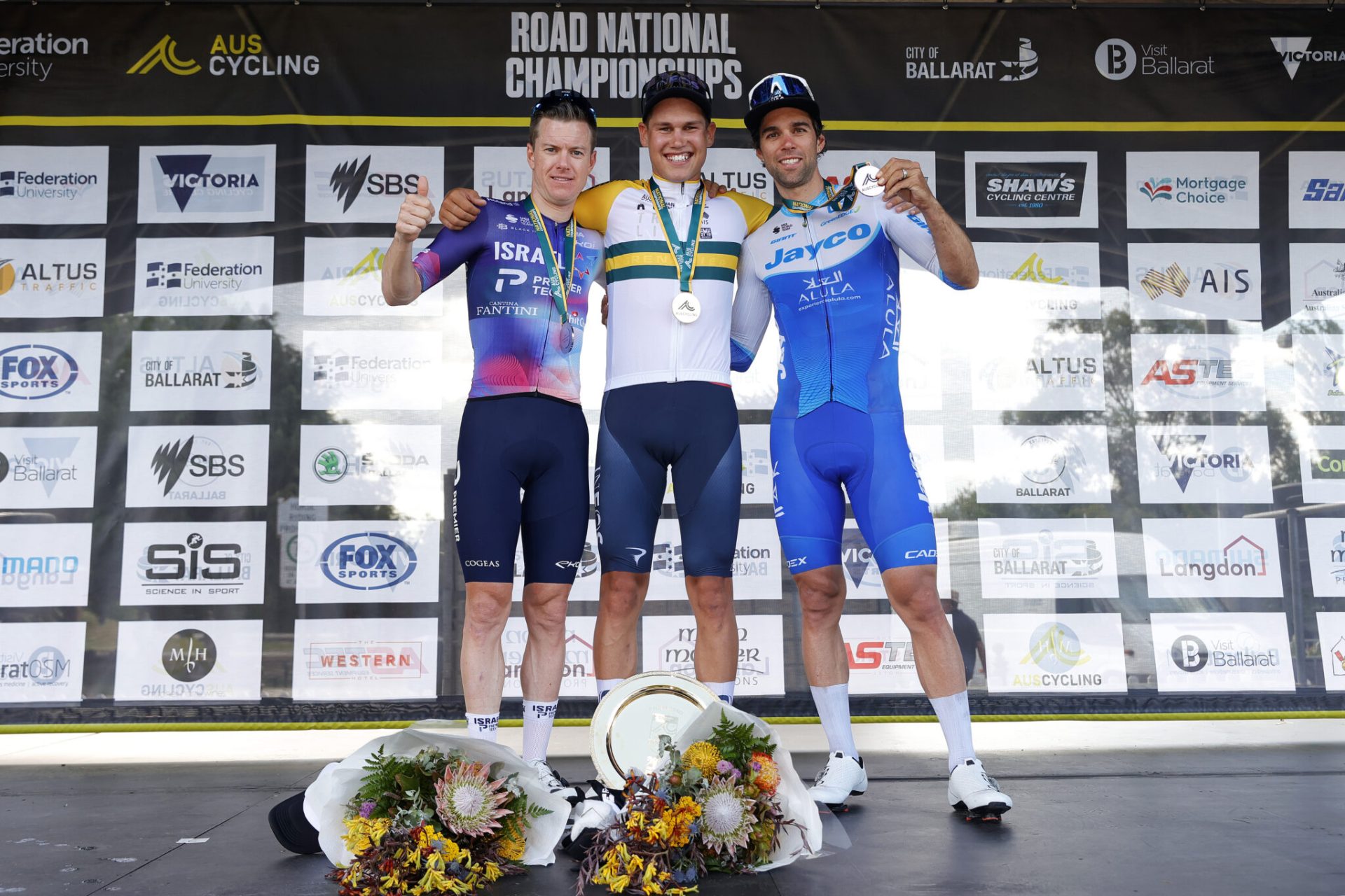 Simon Clarke, Luke Plapp, and Michael Mathews on the podium at Road Nationals, arm in arm, with Plapp in the centre, and Clarke on the left and Matthews on the right. Two bouquets of flowers lie at Plapp's feet.