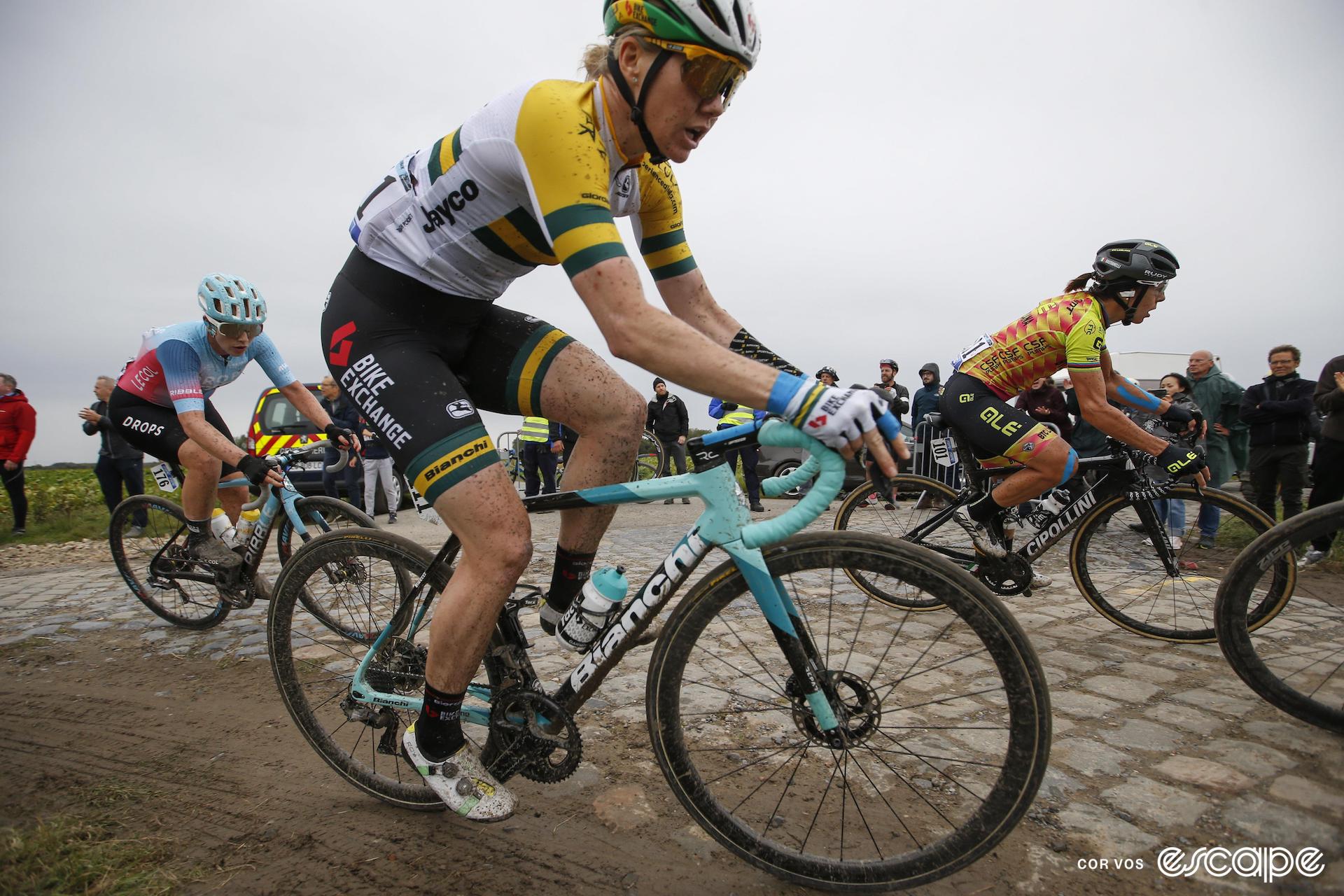 Sarah Roy in the green and gold of Australian road champion rides her Bianchi road bike in the mud beside some cobblestones at Paris-Roubaix.
