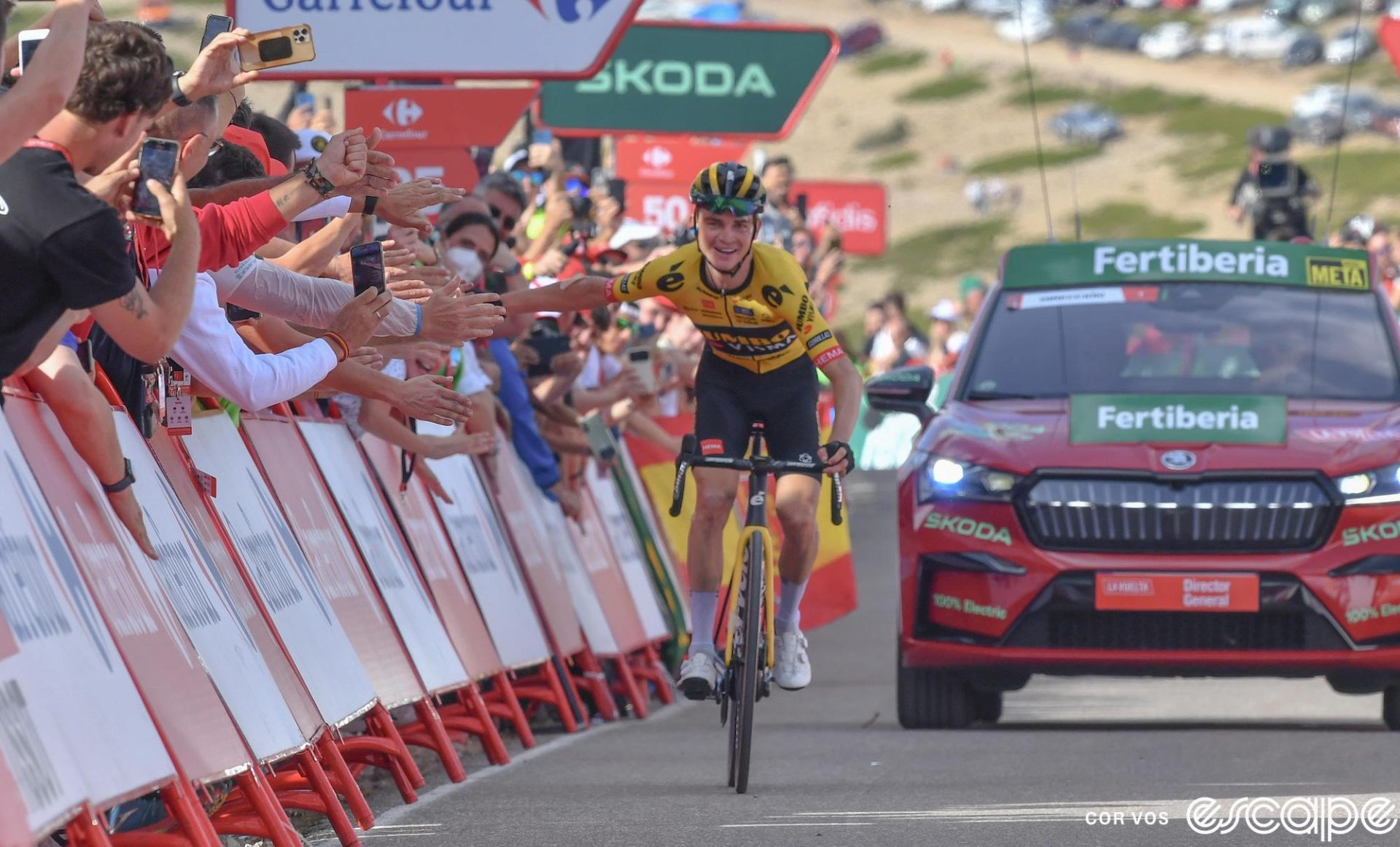 Sepp Kuss rides to the finish on stage 6 of the 2023 Vuelta a España. He's alone, with just the red race official's car behind him, and is riding near the barriers with his arm out to give high-fives to the fans lining the road.