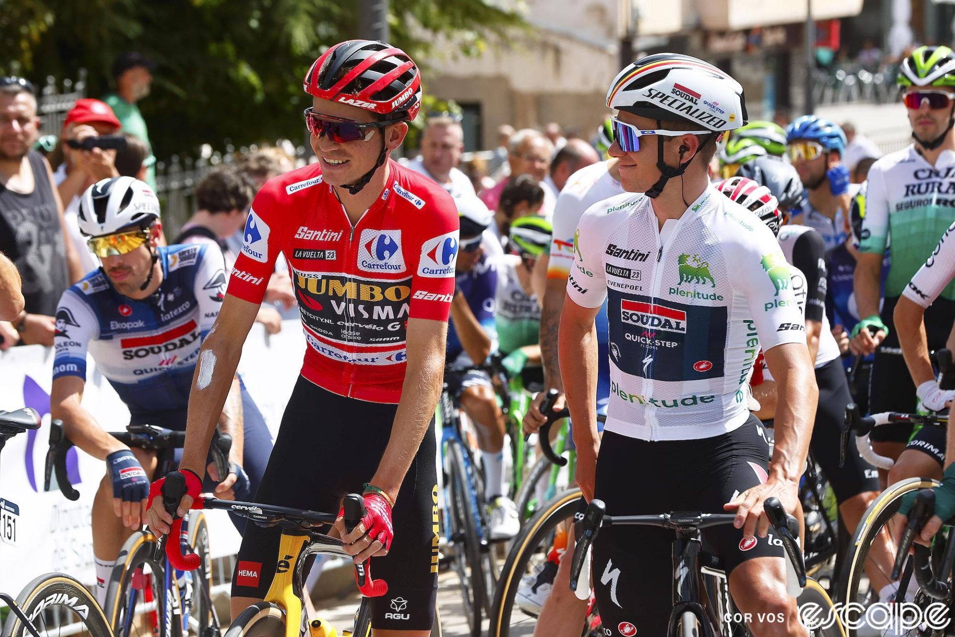 Sepp Kuss and Remco Evenepoel are introduced at the start of stage 12 of the 2023 Vuelta a España. Kuss, on the left, is wearing the red jersey of race leader and has a slight, open-mouth smile on his face as he looks a little to his right. Next to him on his left side is Evenepoel, wearing the white jersey of best young rider. Evenepoel faces Kuss and has a wide, close-mouthed smile on his face.