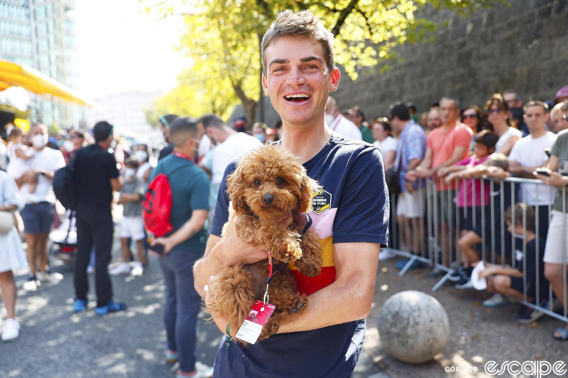 Sepp Kuss holds a small brown Goldendoodle. Kuss is facing the camera, smiling as he holds up the dog, who has its own race credential, labeled "Bimba Kuss." The dog is looking at the camera too, as if to say, am I not a 13/10?