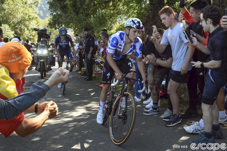 Remco Evenepoel attacks out of the saddle on the La Cruz de Linares climb on stage 18 of the 2023 Vuelta a España. He's wearing the blue polka-dot jersey of best climber, and behind DSM-Firmenich's Max Poole is struggling to hold the wheel as the pair pass a row of fans on the climb.