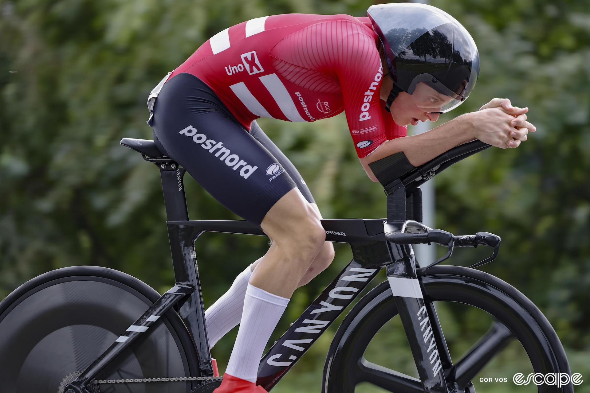 A cyclist rides a time trial in red and black kit with a black aero helmet and white socks.