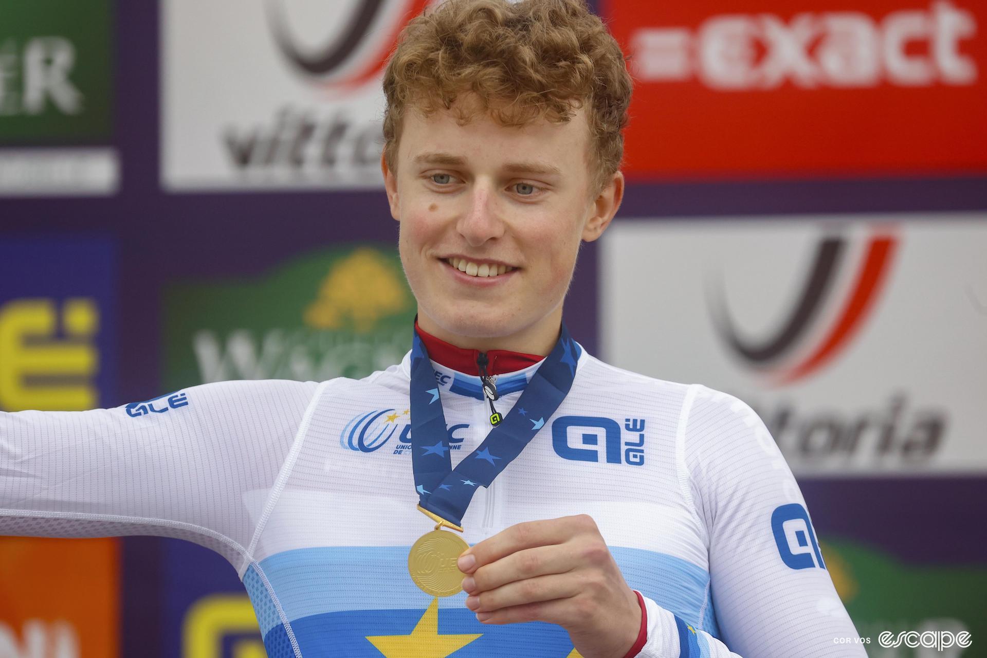 A cyclist in the white and blue jersey of European champion stands on a podium, holding a gold medal that's strung around his neck with a blue strap.