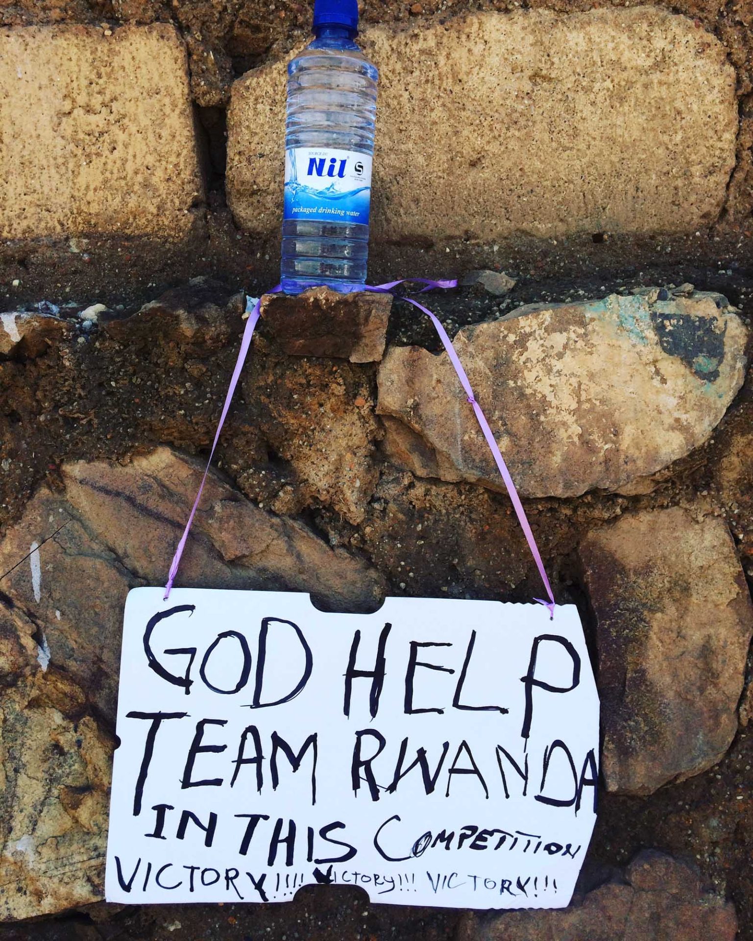 Handwritten sign on a piece of cardboard, reading 'God help Team Rwanda in this competition. Victory! Victory! Victory!'. 