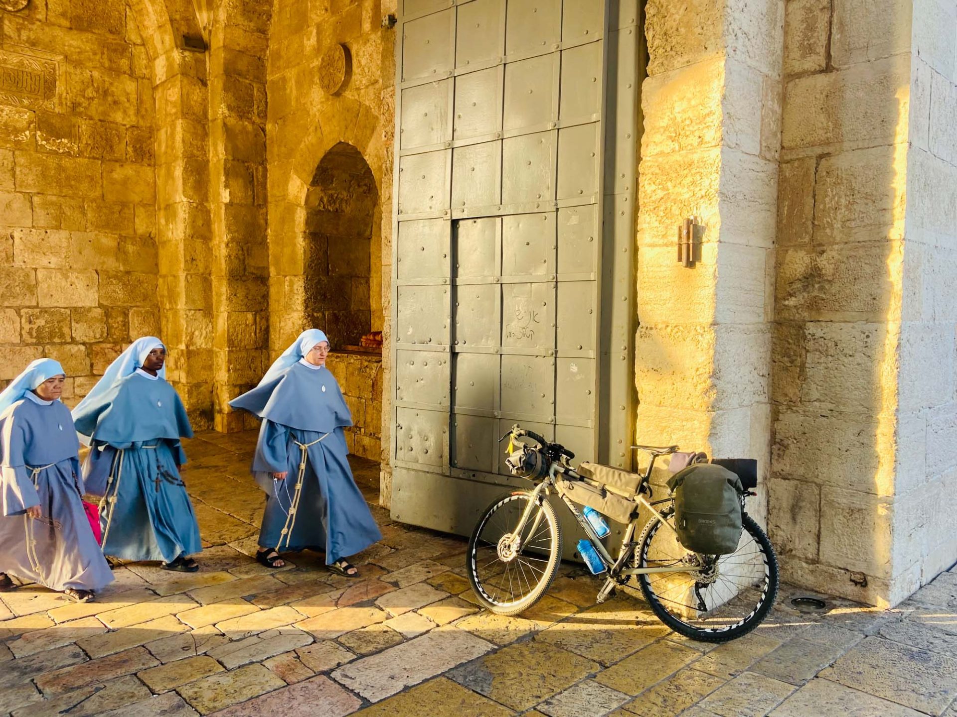 Lazzarin's bike leans against an ancient stone wall, with the sun shining golden rays across the scene. Three nuns in habits walk past. 