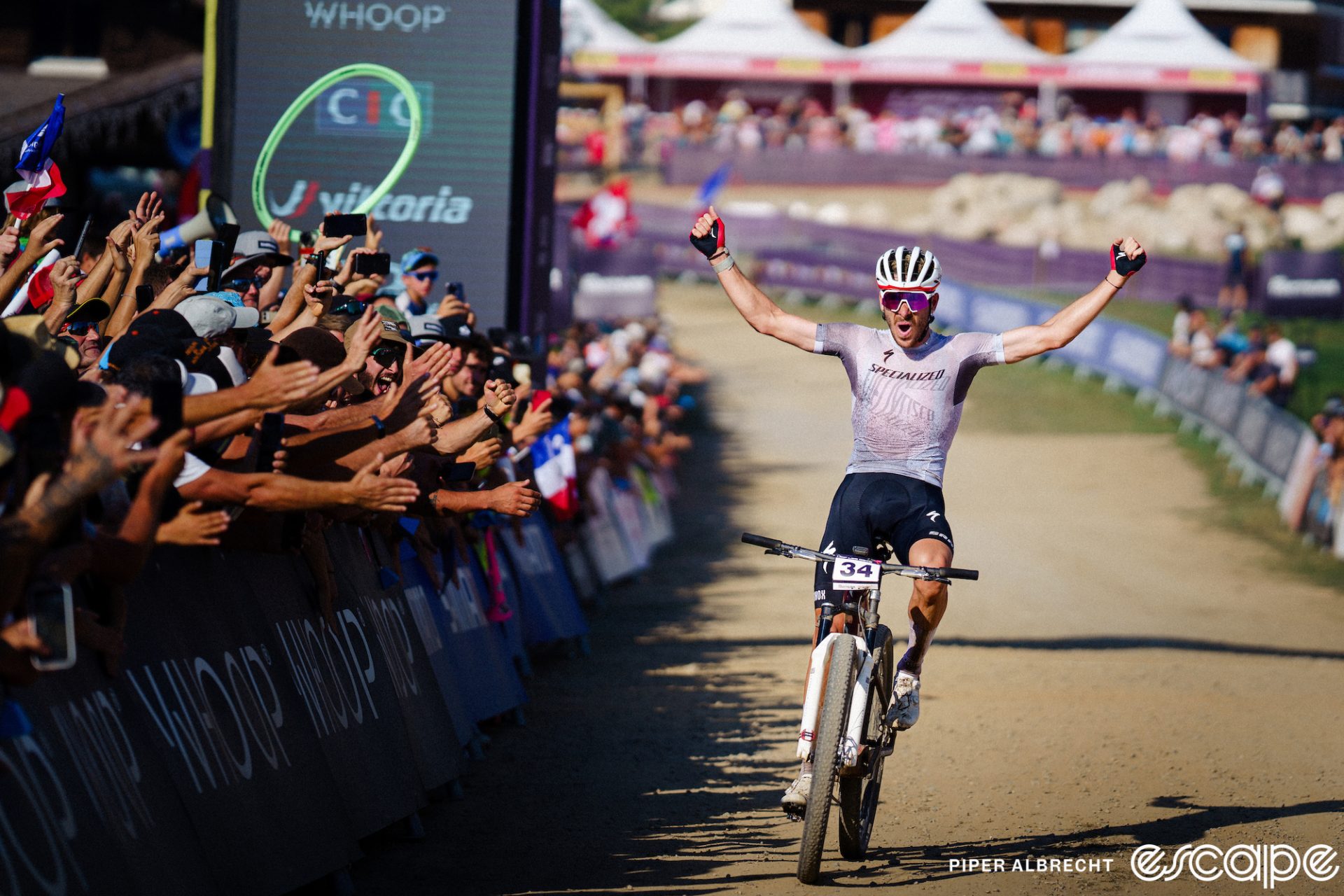 Victor Koretzky crosses the finish line to win the Les Gets round of the 2023 World Cup XCO series. He's all alone, arms in the air, as a large crowd cheers him on.