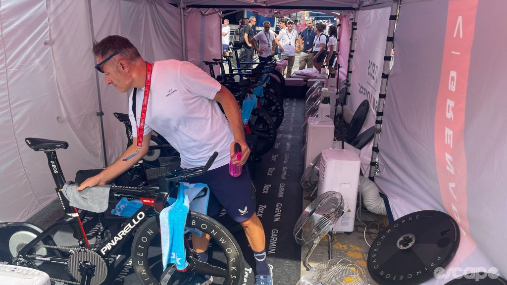 The photo shows inside the Ineos Grenadiers cool tent. We can see the team bikes on stationary trainers with fans and air conditioning units in front of each ride.