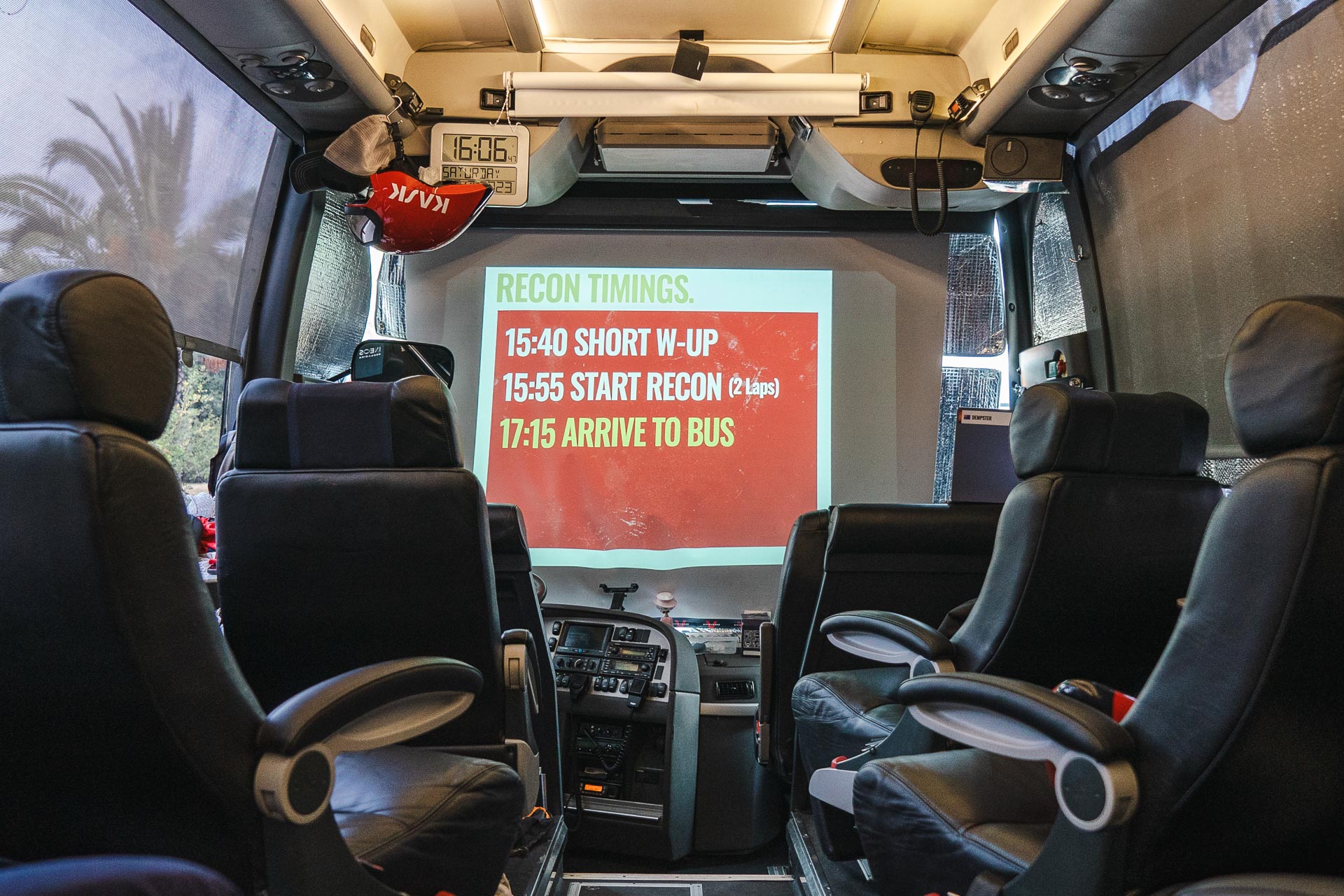The image shows a photo from inside the Ineos Grenadiers team bus with a projector displaying the timetable for the day on a pull-down screen. The time table reads: Recon times. 15:40 Short warm-up. 15:55 Start Recon (2 laps). 17:15 Arrive to bus. A clock on the bus shows it is currently 16:40