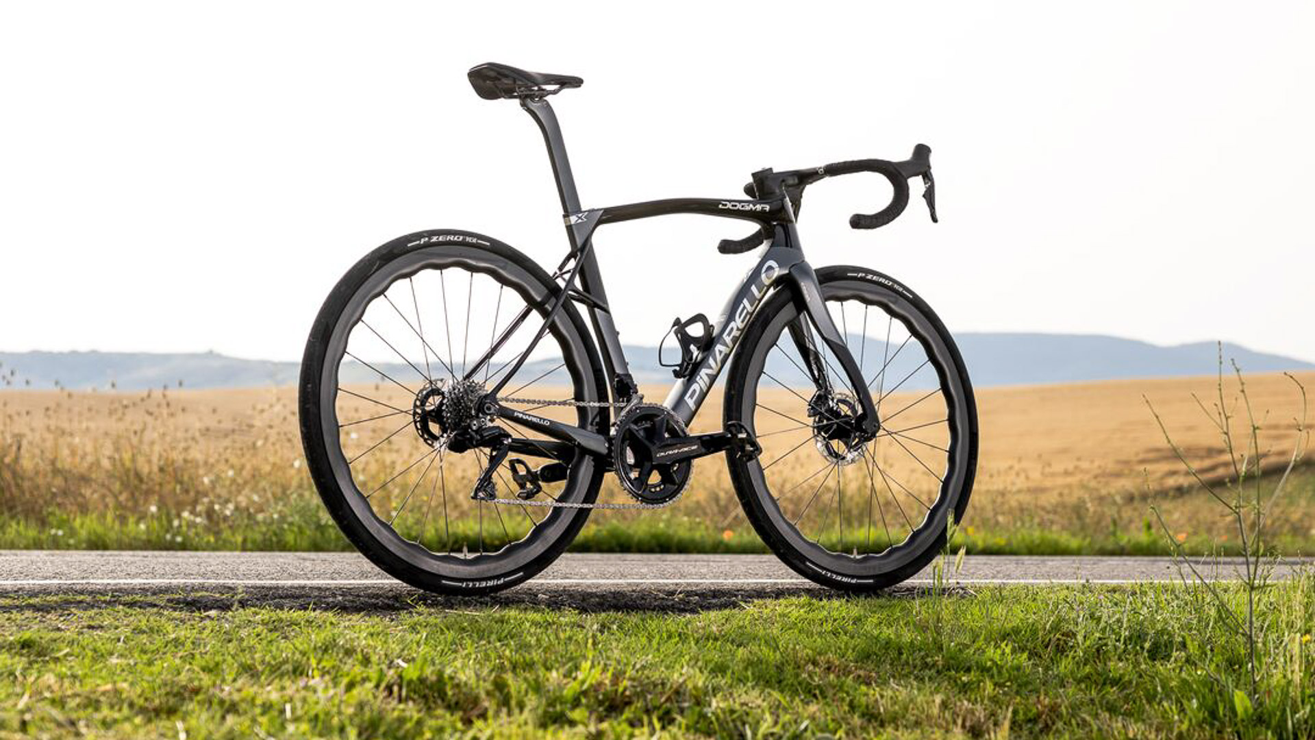 Pinarello's new Dogma X endurance bike. The bike is pictured along a road side in a rural setting with rolling hills in the far background, golden fields just behind the bike with the bike standing seemingly unaided on the tarmac edge by a grassy border. The image is side on and slightly from the rear to showcase the X-Stay seat stay design.