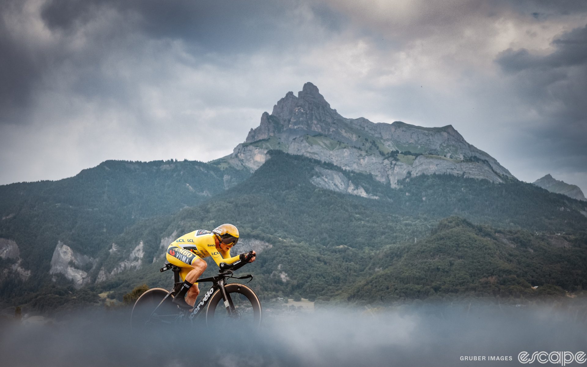 Jonas Vingegaard time trials at the 2023 Tour de France. He's in an exceptionally aero position, shown alone on a backdrop of a rugged mountain behind him with a steep, rocky peak, and the foreground is slightly blurred to create a moody feel.
