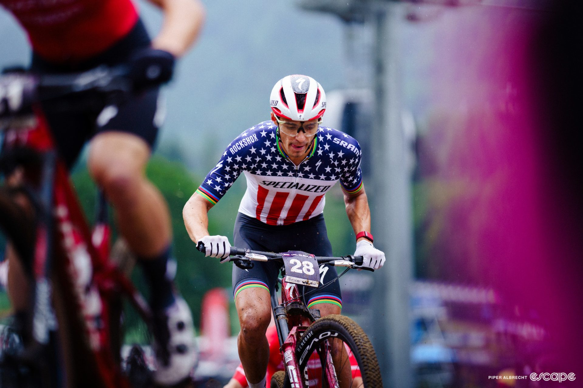 Chris Blevins races in a World Cup in Europe. He's focusing on a technical bit of trail, looking down, and is wearing the stars-and-strips jersey of national champion, with rainbow bands at his sleeves and short cuffs to honor his short-track World Championship in 2021.