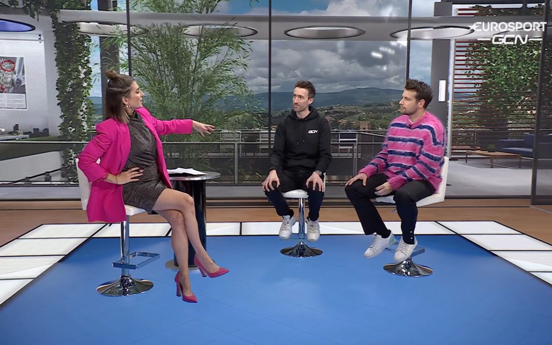 Orla Chennaoui, Dan Lloyd, and Adam Blythe sit in chairs on the GCN set. Adam Blythe is wearing the world's ugliest jumper, a horrible mix of pink and purple.